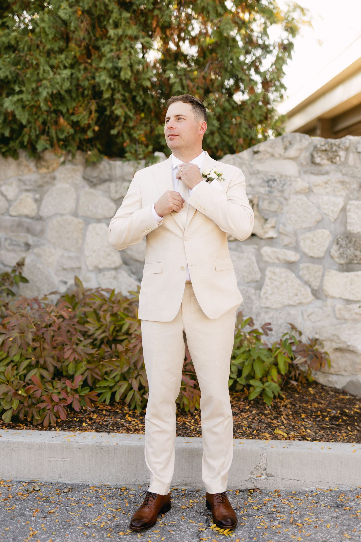 A person in a cream suit adjusts their tie against a stone wall backdrop.