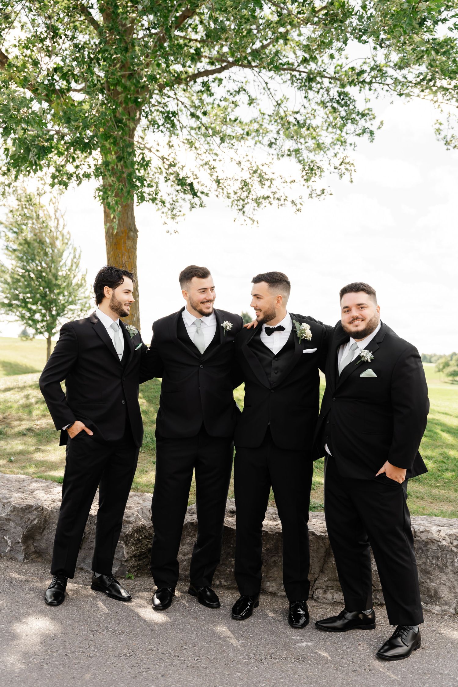 Groomsmen in black tuxedos pose casually against outdoor backdrop with trees.