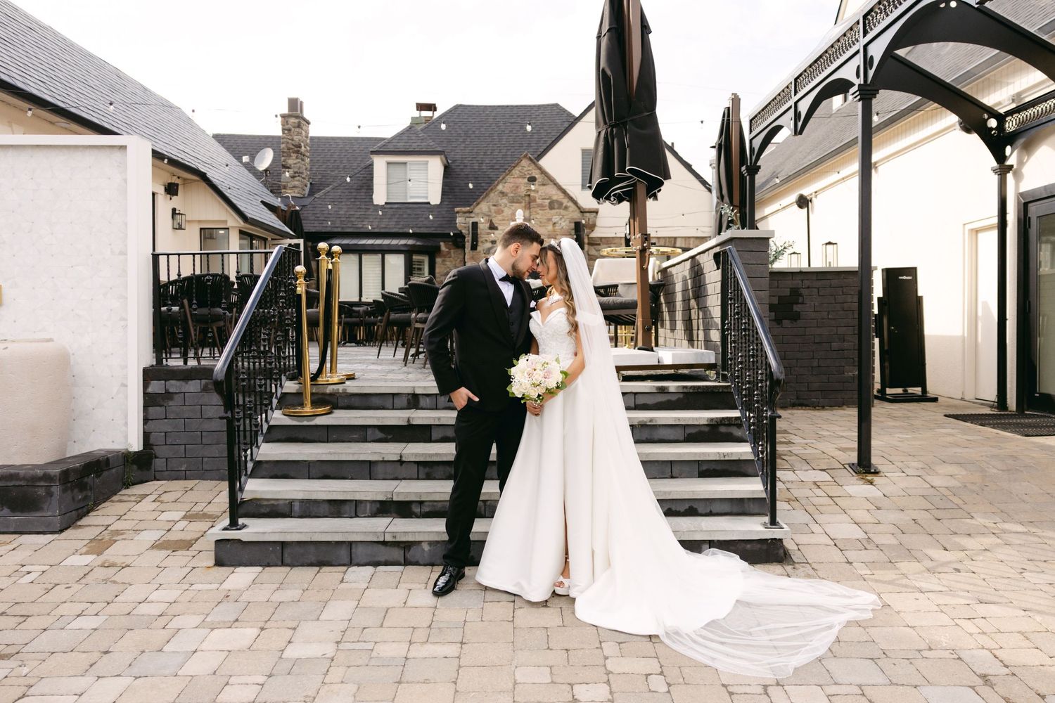 Elegant wedding couple poses on stone patio with flowing white dress and veil against modern architecture.