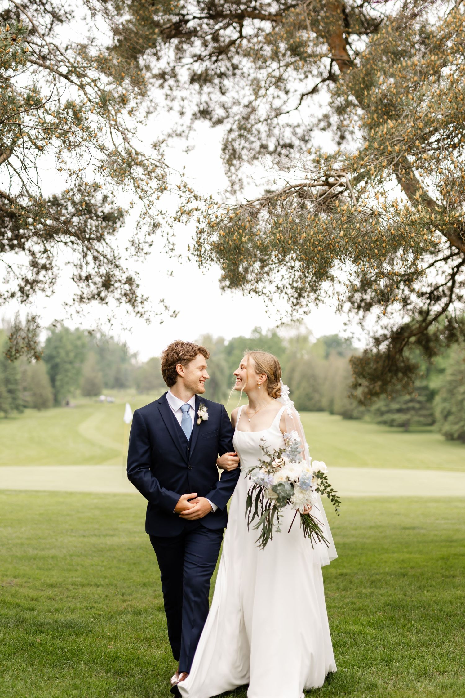 Wedding couple strolls on lush golf course under overhanging pine branches.