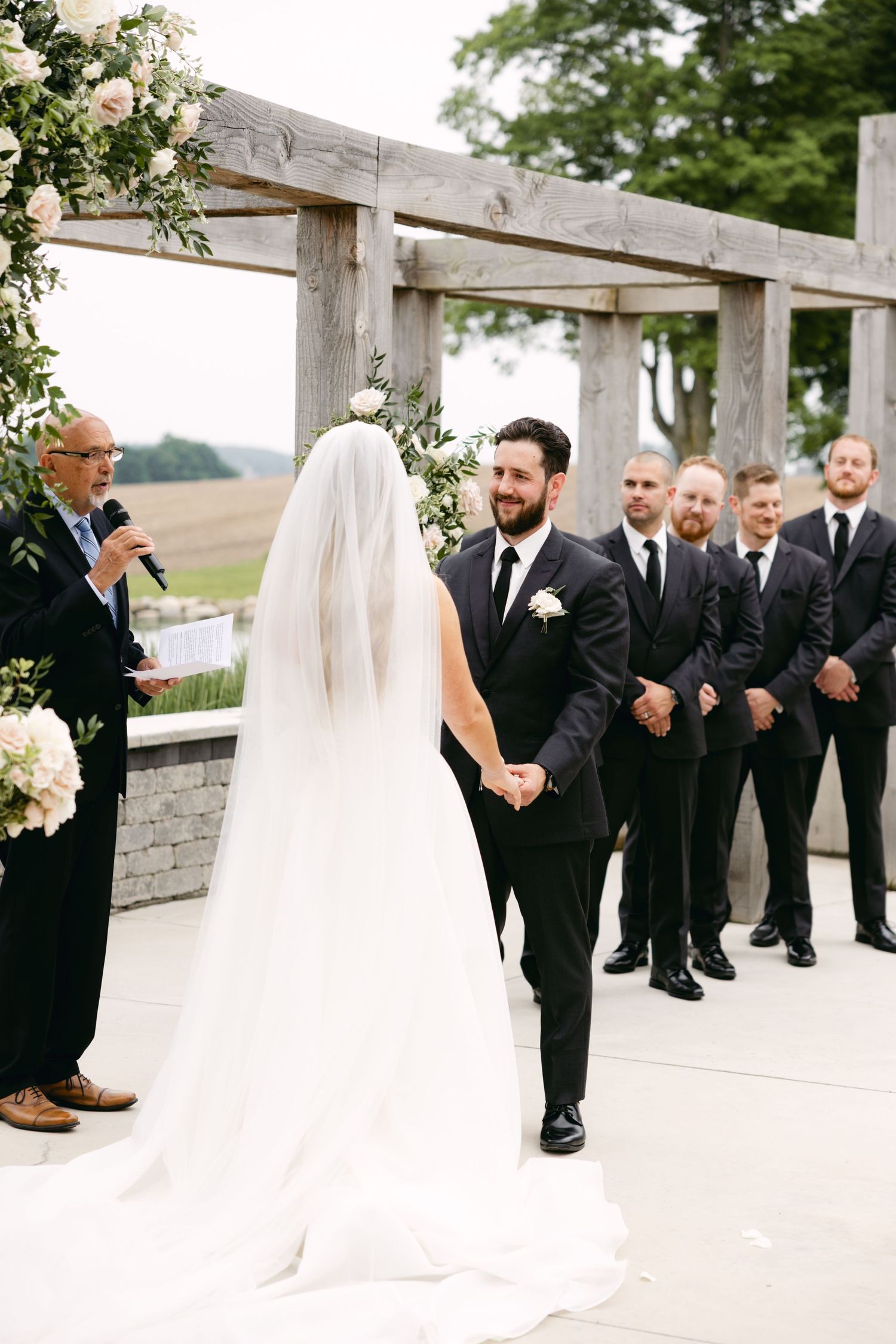 Outdoor wedding ceremony with attendants in black suits standing near a stone wall and wooden pergola.