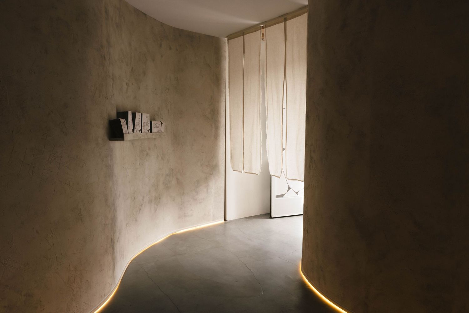 Minimalist curved corridor with warm LED strip lighting, textured plaster walls, and draped fabric doorway in neutral tones.