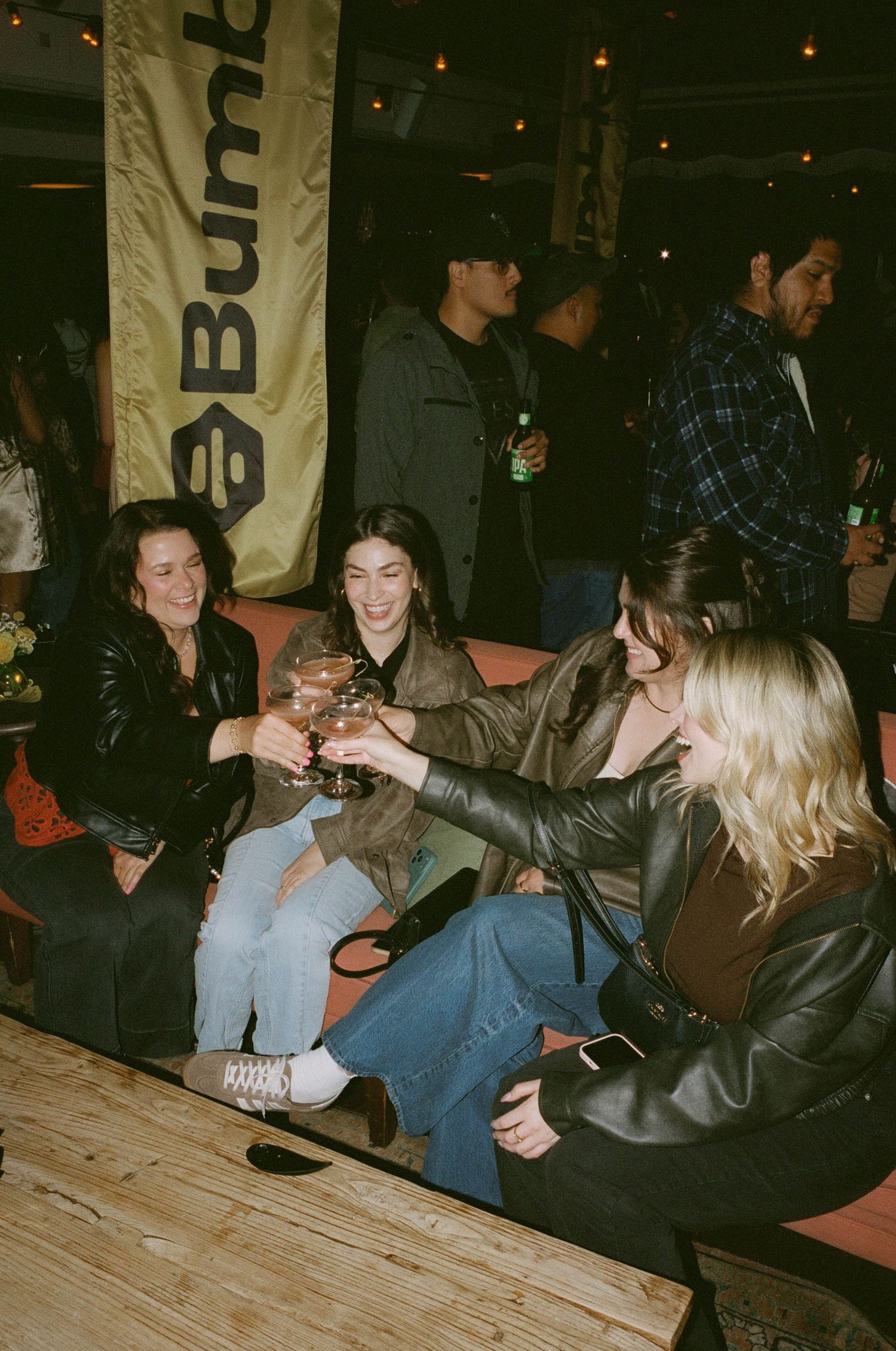 Four women toast drinks at a Bumble event, wearing leather jackets, gathered on wooden seating in a dimly lit venue.
