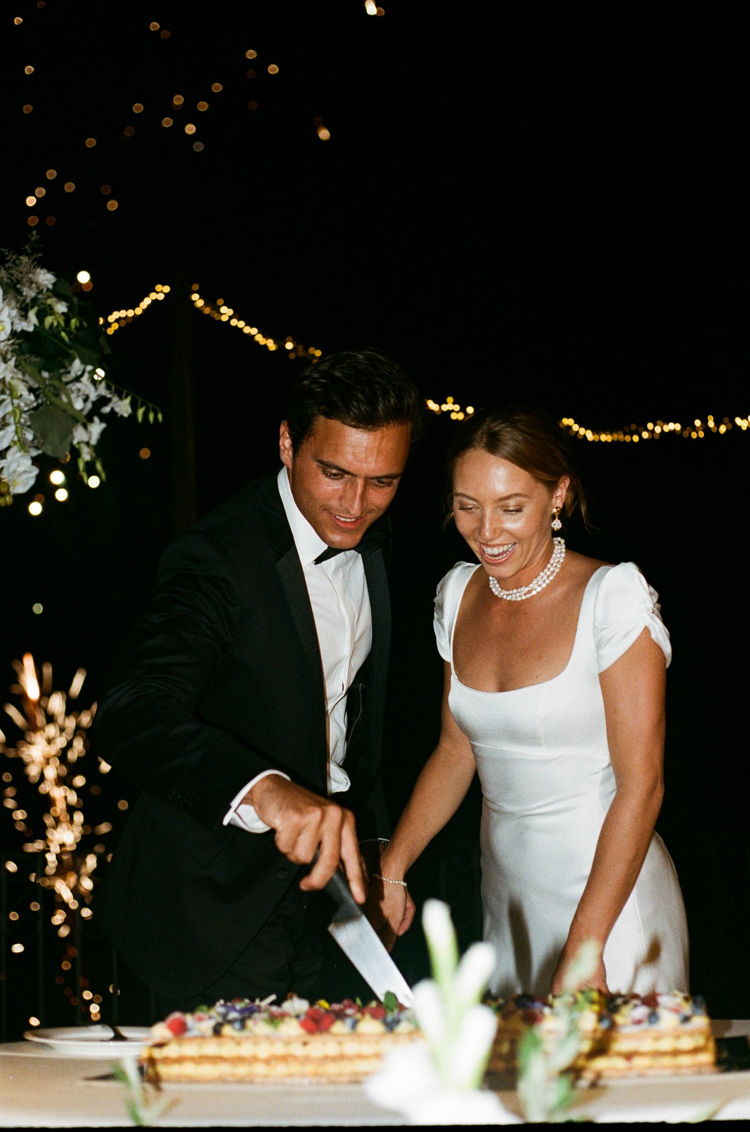 Couple cutting their wedding cake together with sparklers in the background.