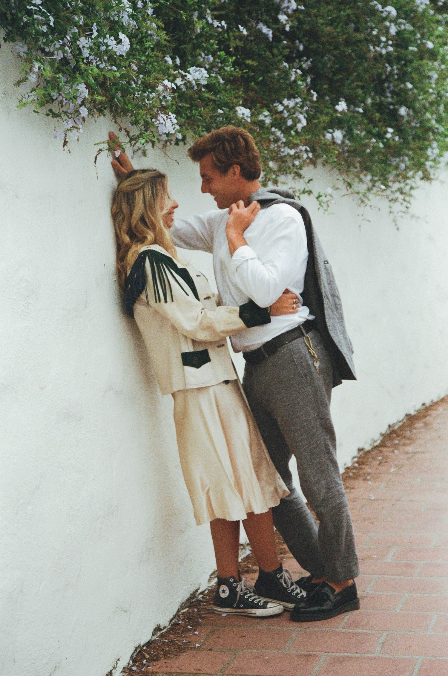 Two people in casual attire lean against a white wall while sharing an intimate moment on a brick pathway.