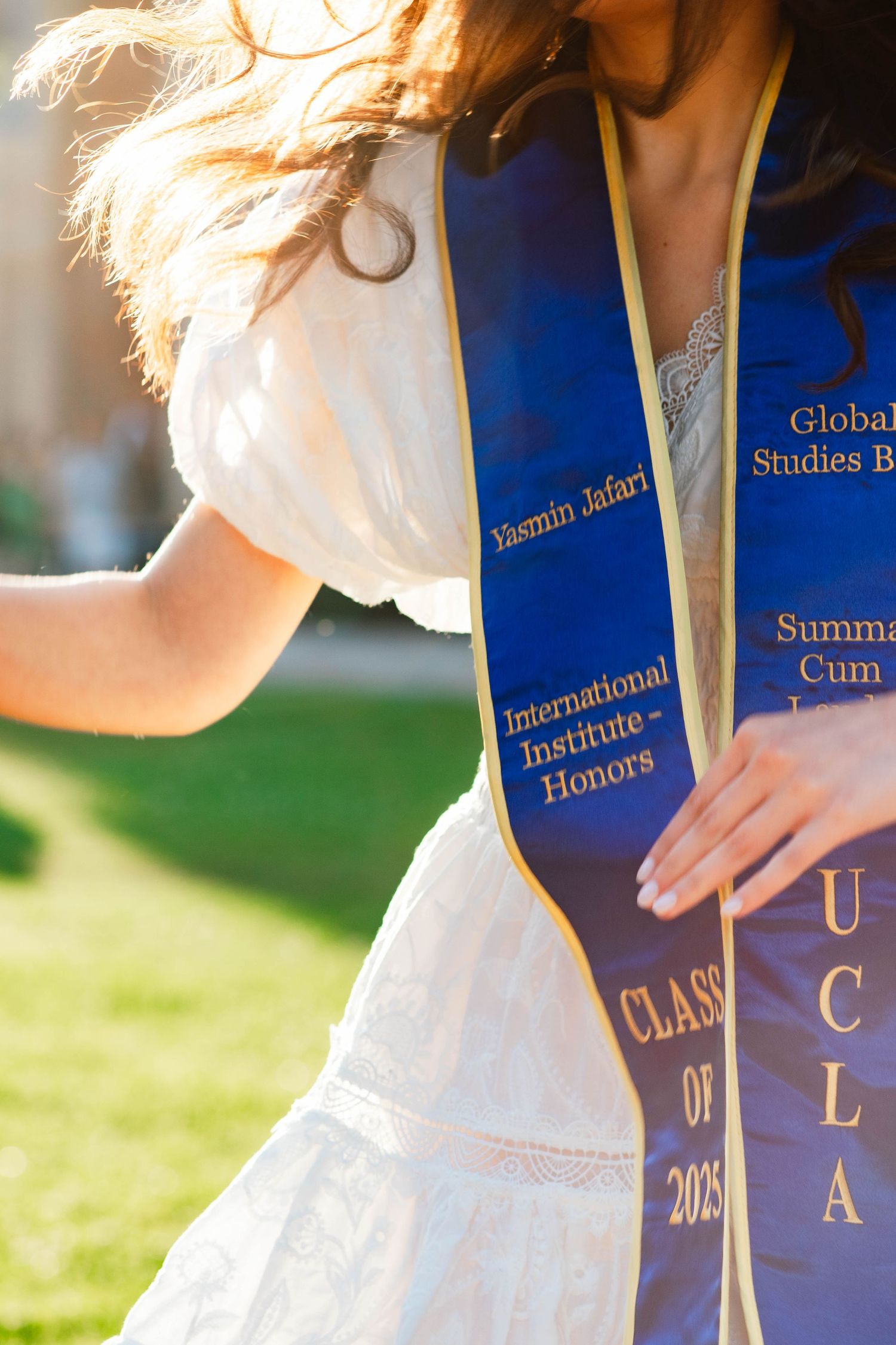 Graduate in white dress poses with blue graduation stoles and diplomas against outdoor background.