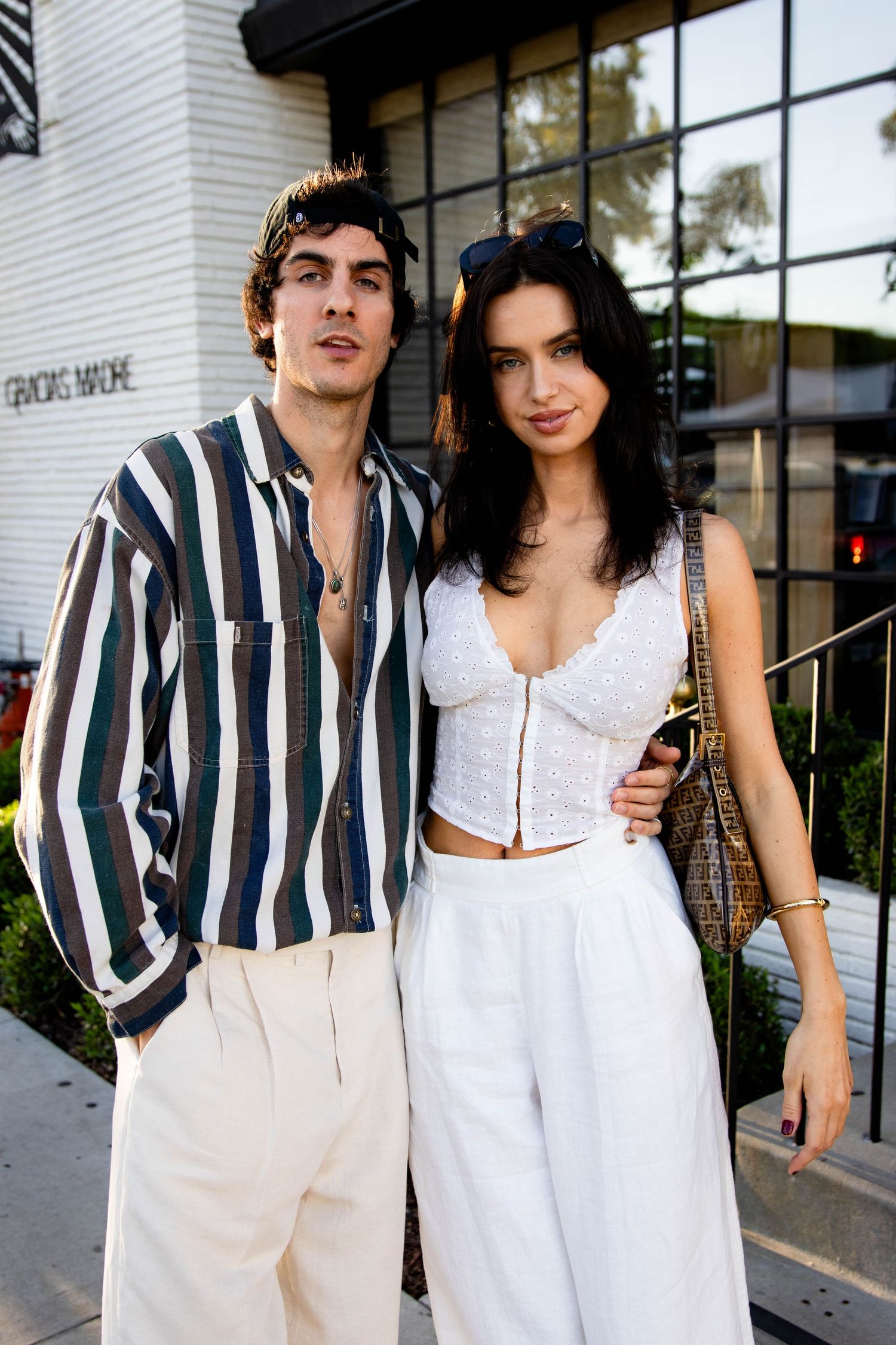 A fashionable couple in coordinated outfits poses outside a building, wearing striped shirt and white ensemble.