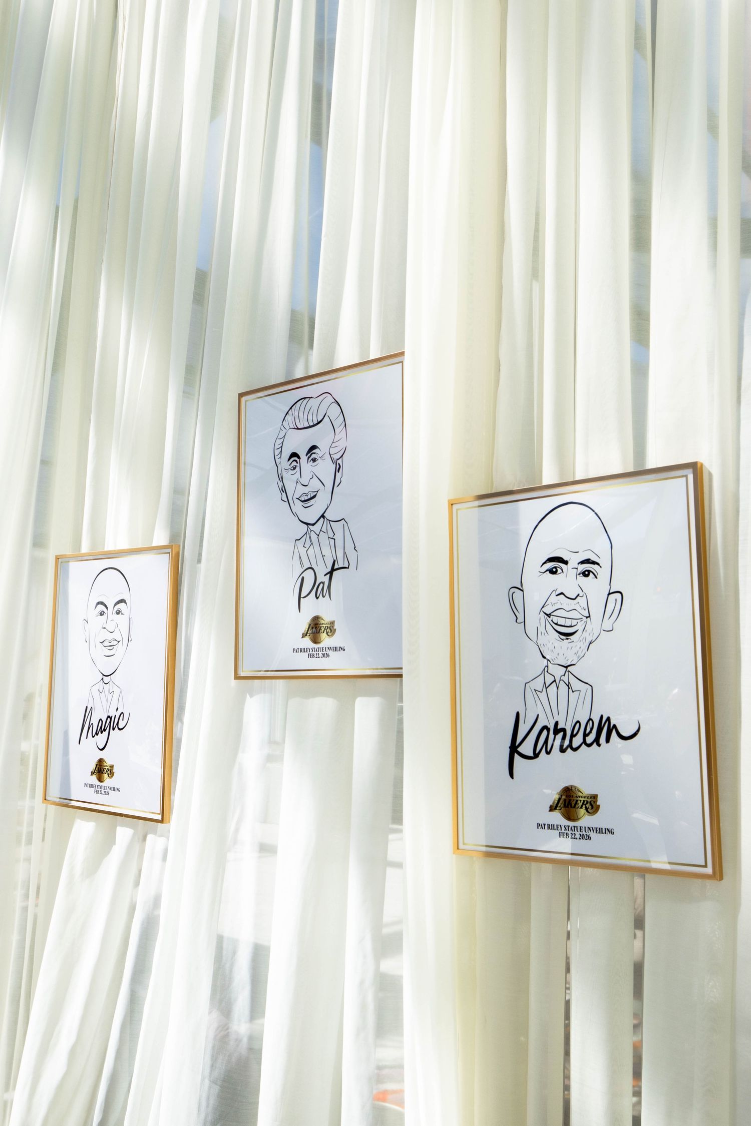 Framed caricature drawings of Magic, Pat, and Kareem displayed against sheer white curtains at a Lakers event.
