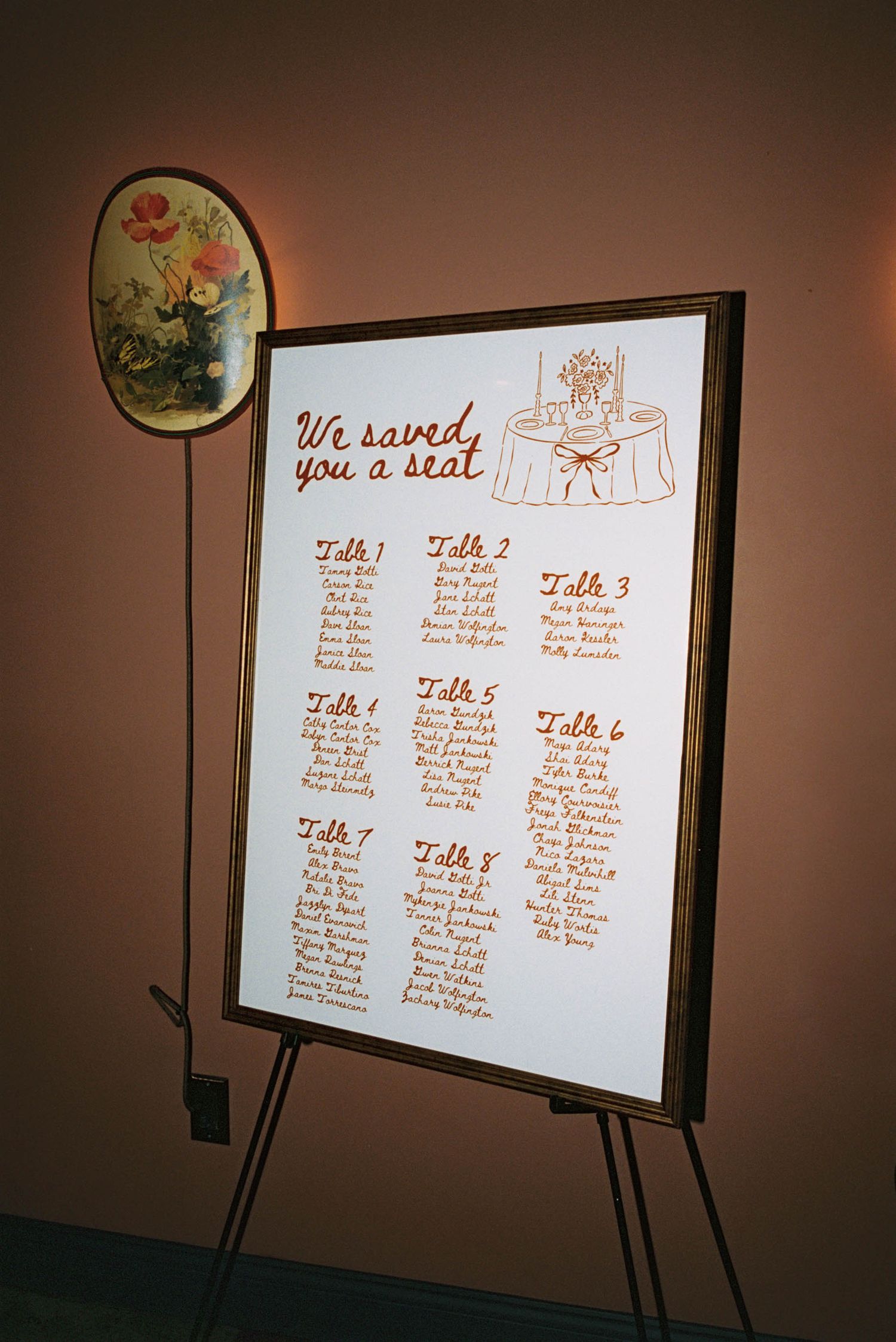 A wedding seating chart display on an easel shows table assignments under the text 'We saved you a seat' with floral decor.