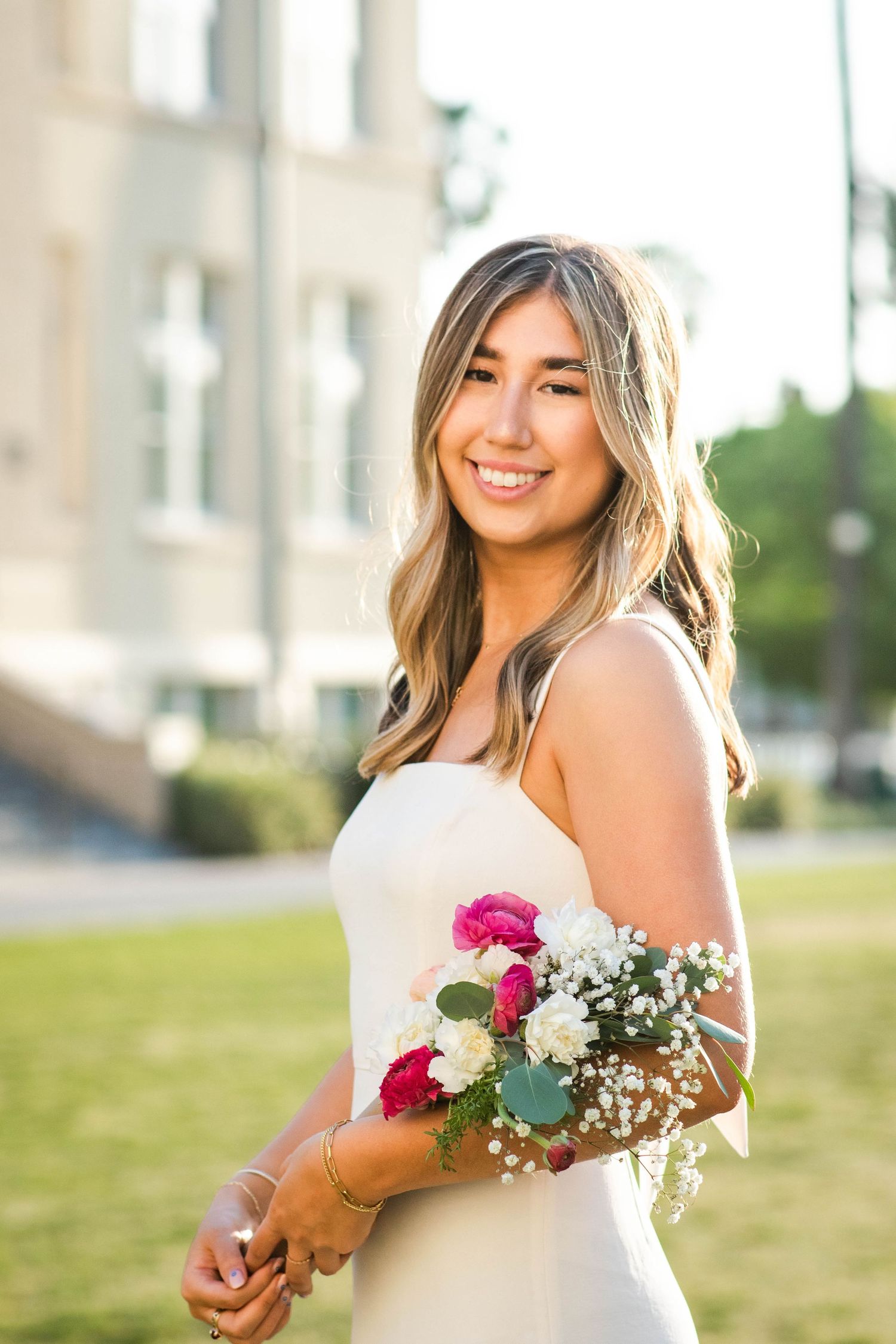 Graduate in white dress holding pink flowers during outdoor photo session.