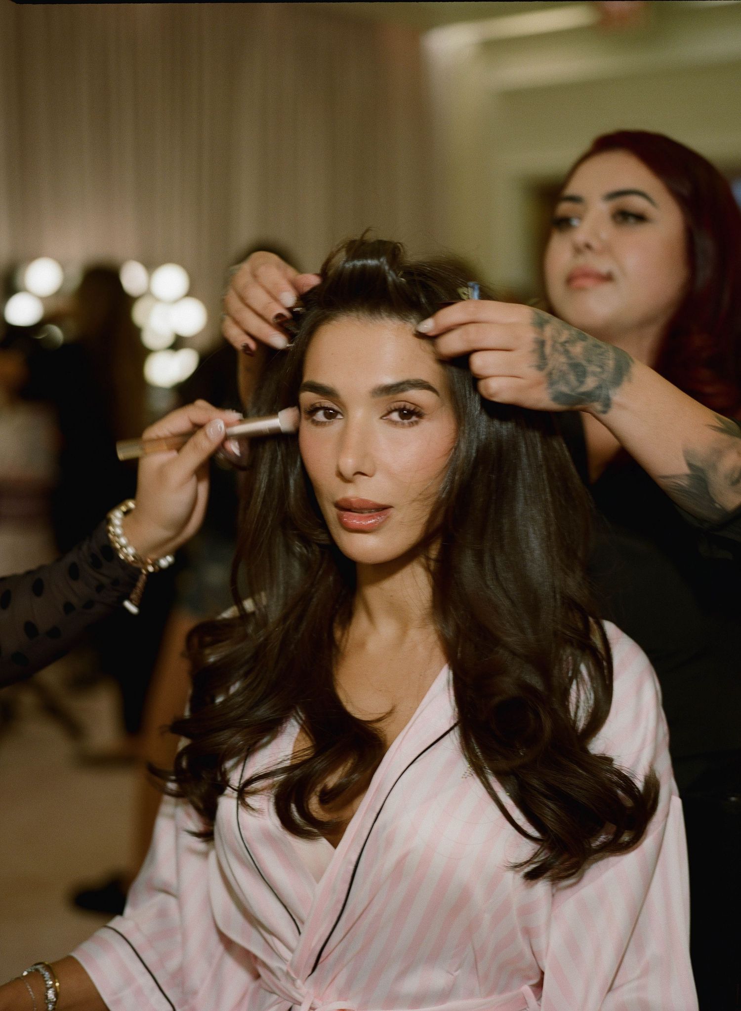 Woman with long dark wavy hair in a pink striped satin robe getting makeup and hair styling backstage under bright vanity lights.