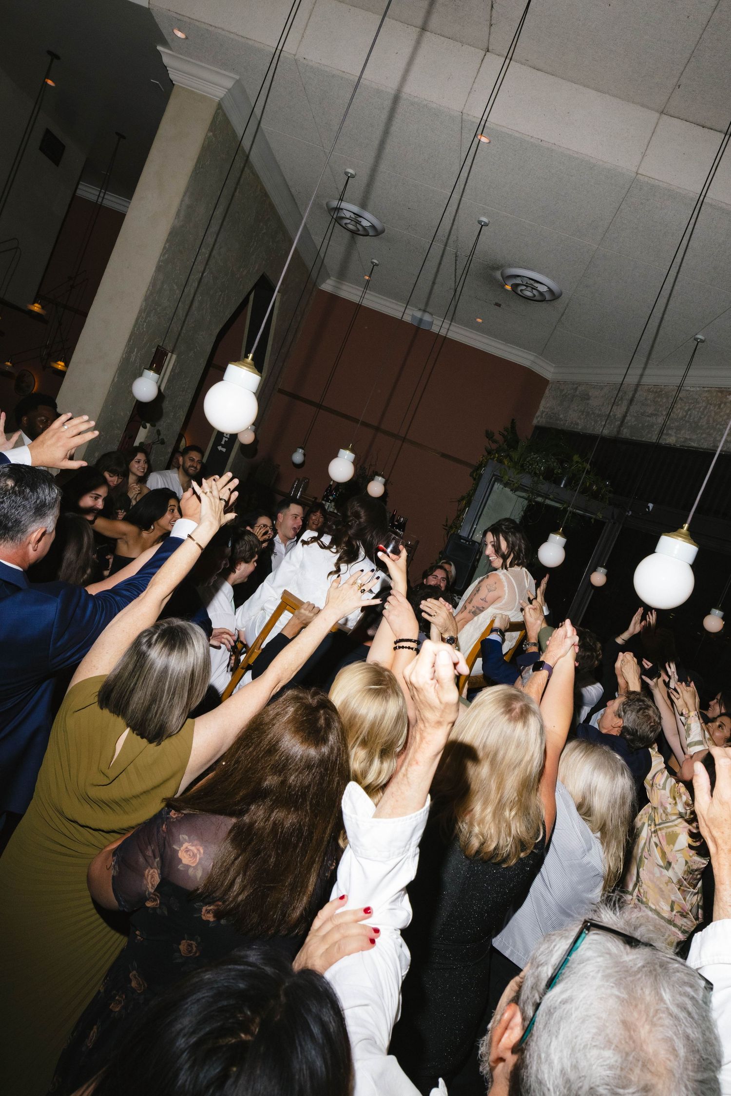 Crowd of party guests with raised hands dancing under hanging light bulbs.