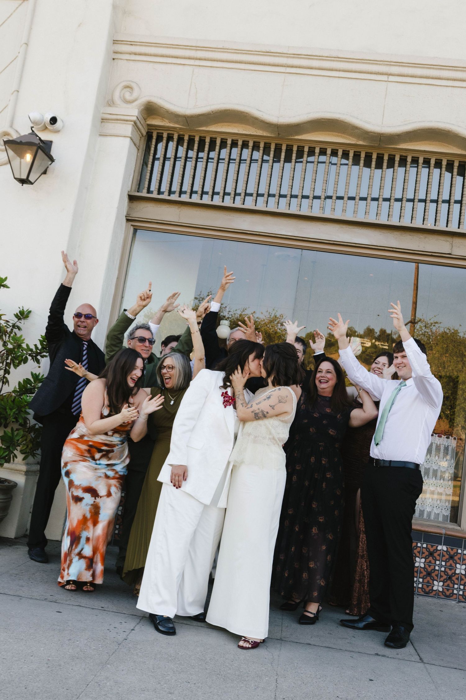 Wedding party celebrating outside building with raised arms and joyful expressions.