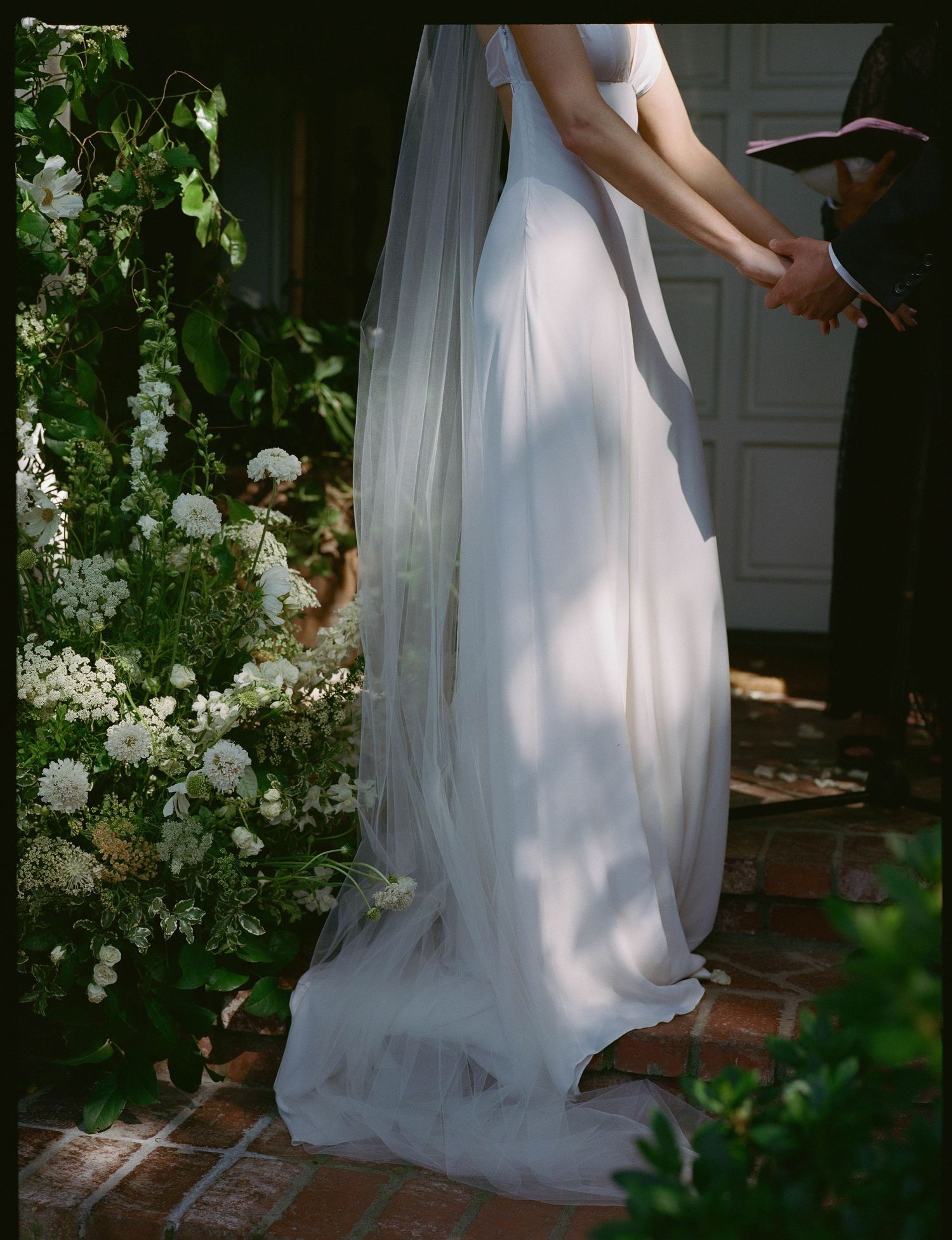Bride in flowing white wedding gown and veil holding hands with groom, surrounded by lush greenery and white floral arrangements.