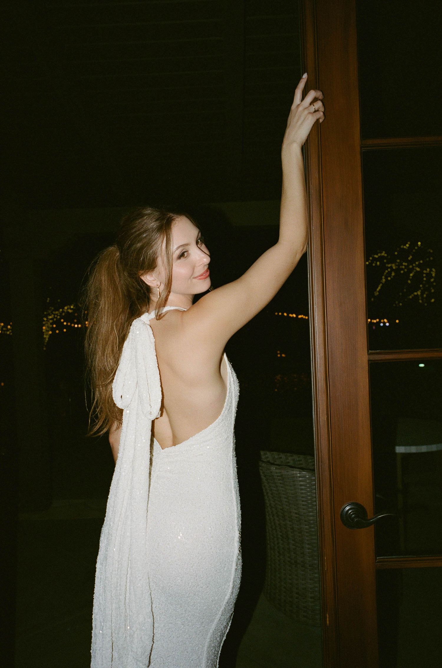 Woman in a white sequined backless gown with a long ponytail glances over her shoulder while holding a wooden door frame at night.