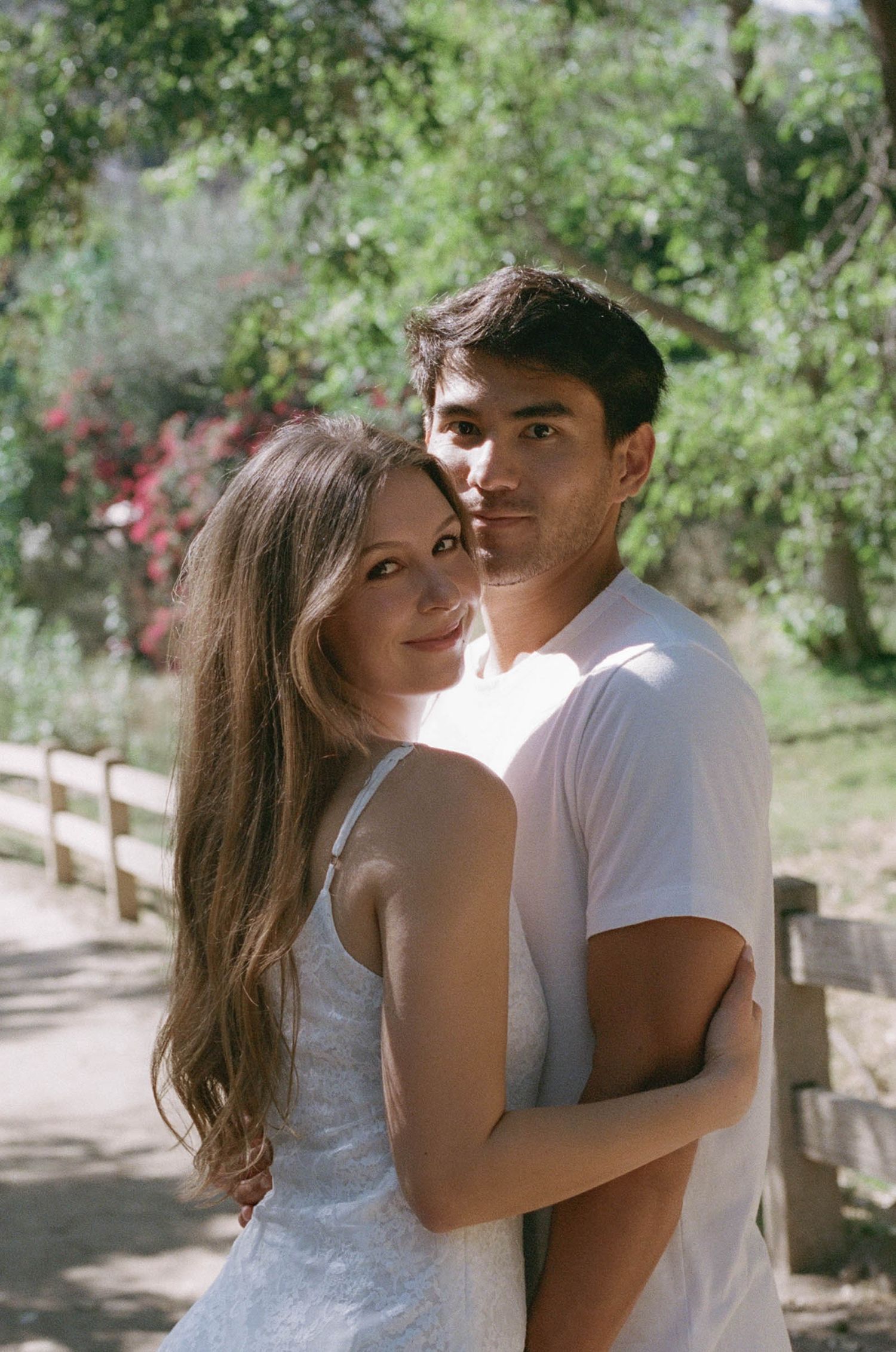A young couple embracing outdoors on a sunny day, surrounded by lush green trees and a wooden fence.