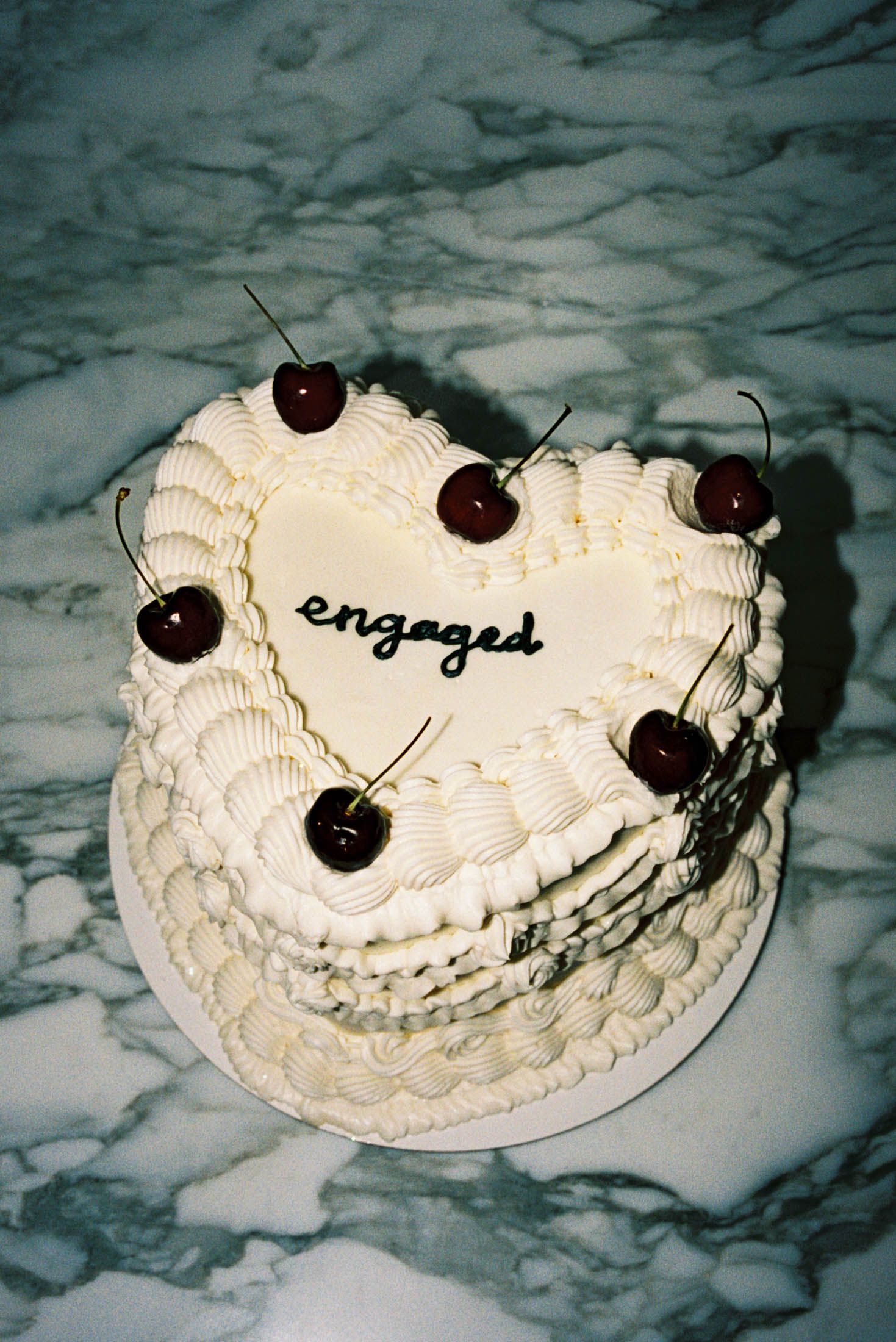A white heart-shaped cake decorated with cherries and the word 'engaged' written in script on top against a marble background.