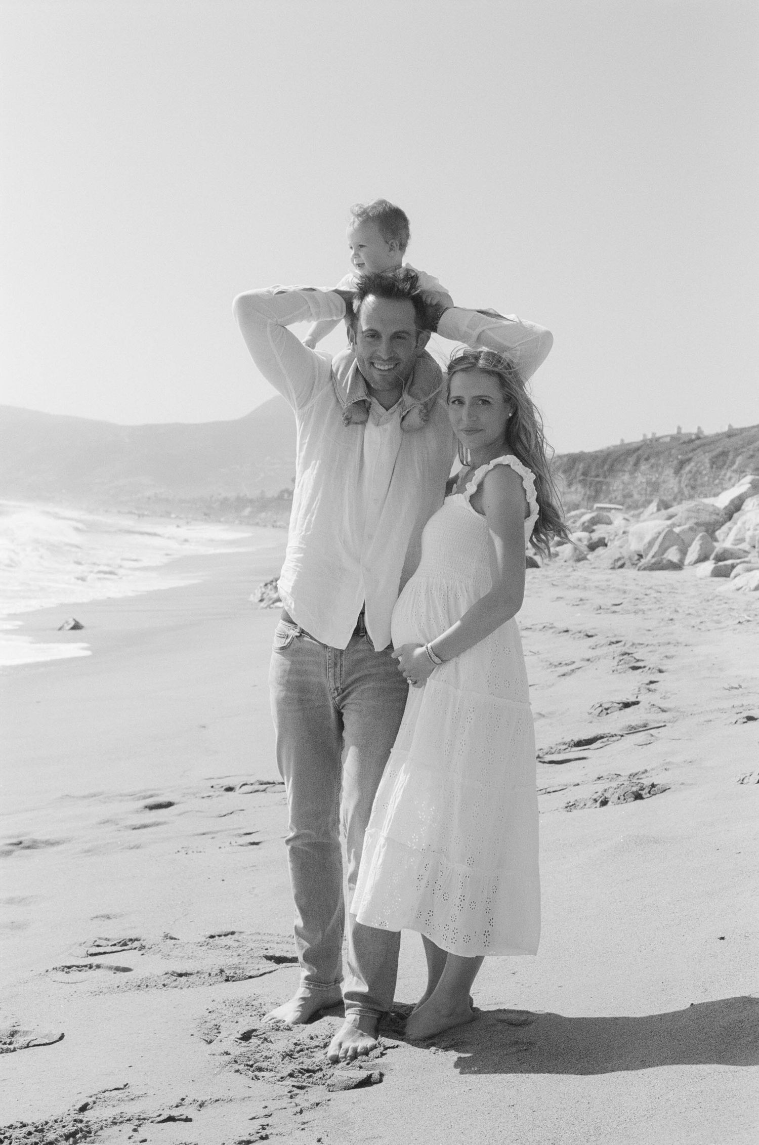 Black and white beach photo of a smiling couple with a young child on the man's shoulders, ocean waves in the background.