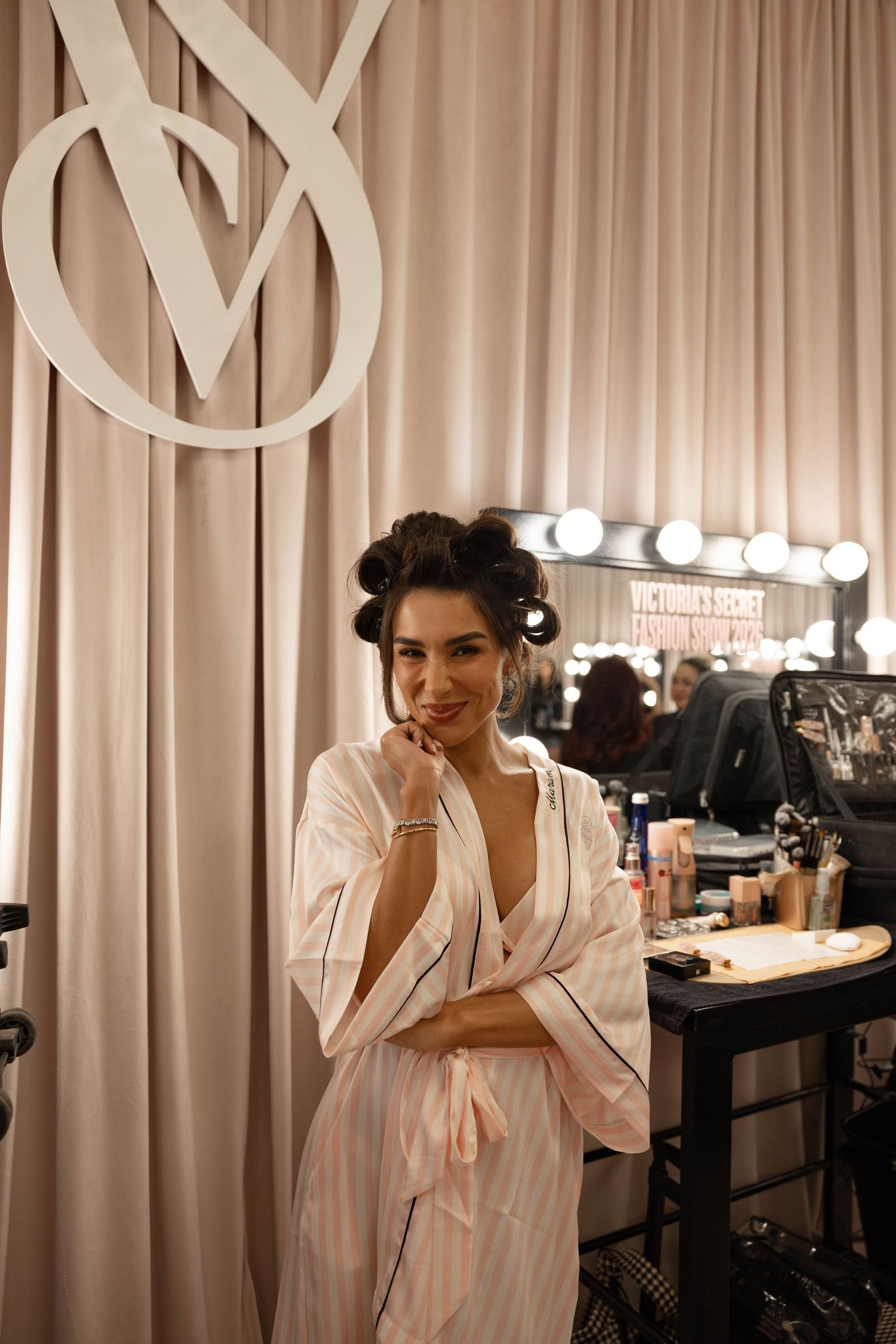 Woman in a pink striped satin robe and hair rollers poses backstage beside a Victoria's Secret logo and a lighted makeup mirror.