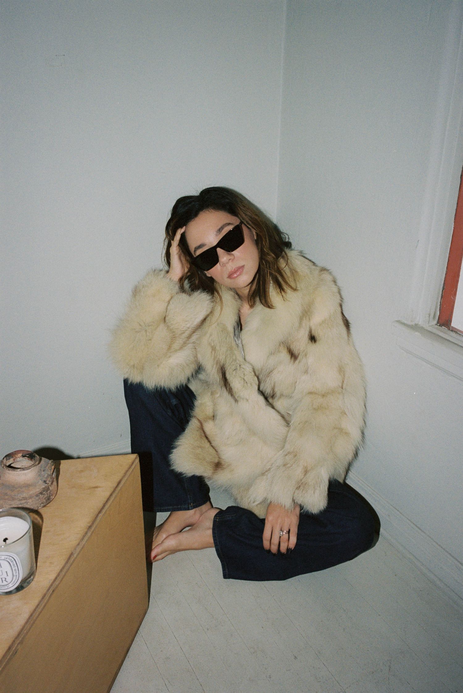 Person in cream fur coat and dark pants sits on floor in minimalist white room across multiple candid shots.