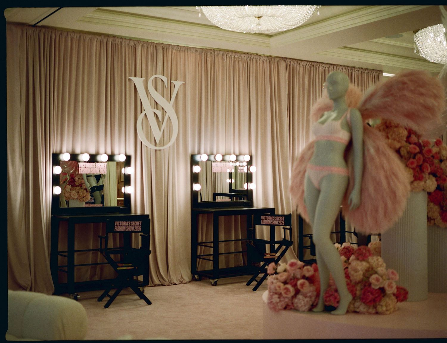 Backstage beauty room with lighted vanity mirrors, makeup chairs, and a mannequin in pink lingerie with feathered wings beside rose arrangem