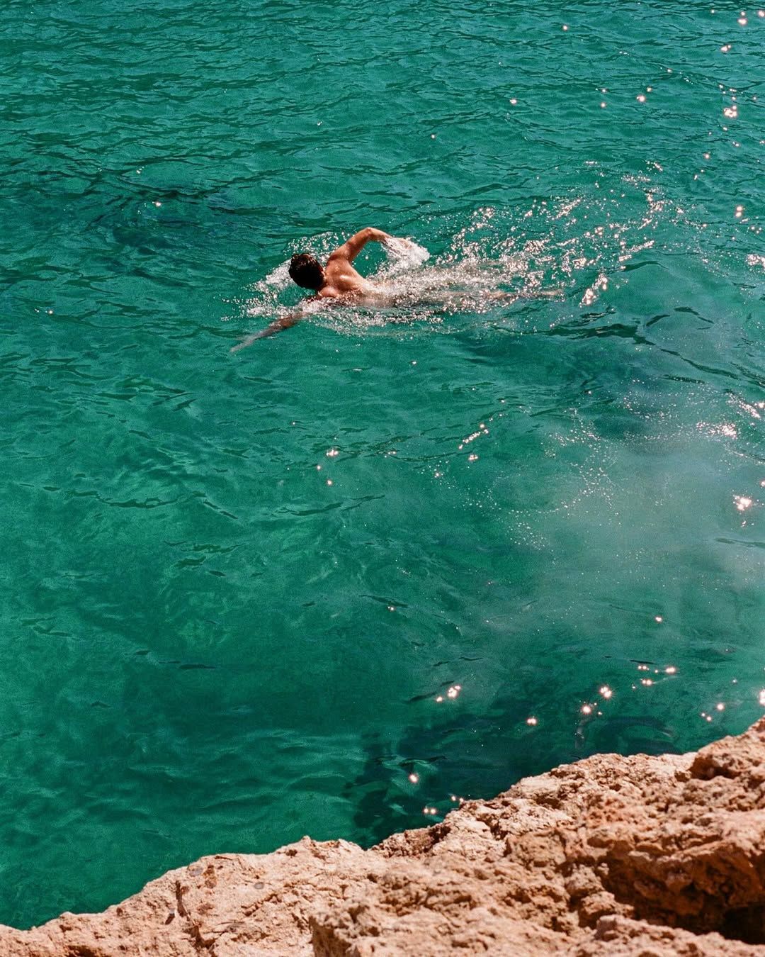 Turquoise Mediterranean waters lap against rocky limestone cliffs on a sunny summer day.