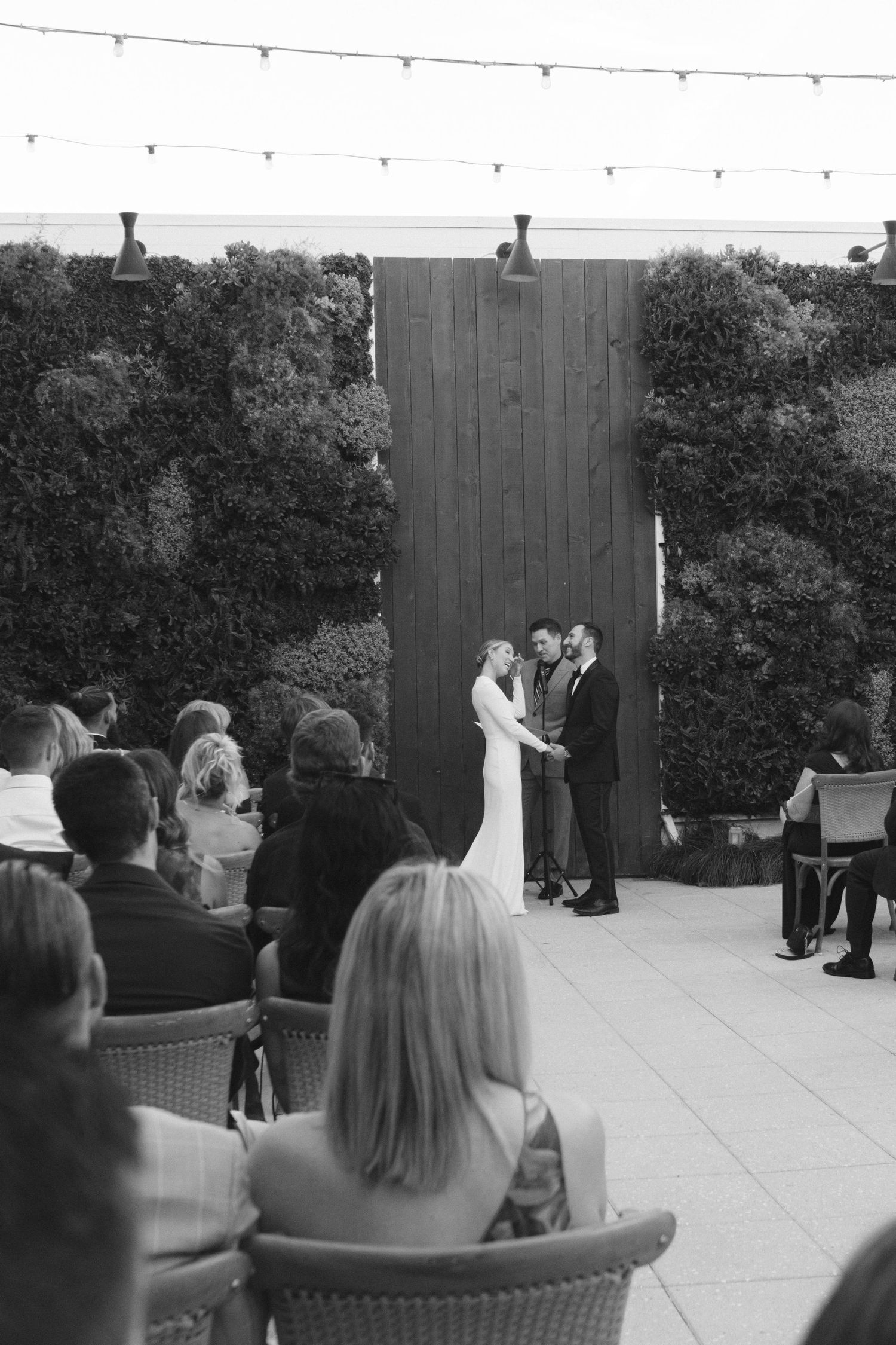 Outdoor wedding ceremony with wooden backdrop and string lights as guests watch the couple exchange vows.