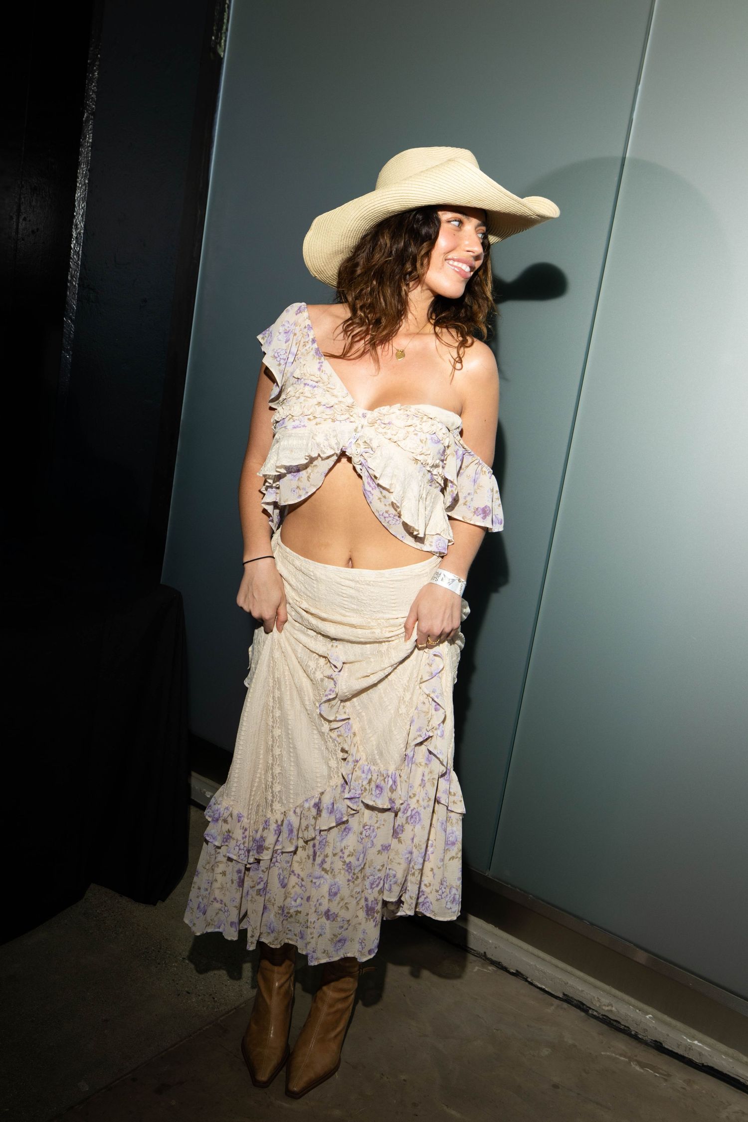A person in a cream-colored cowboy hat and floral prairie dress with brown boots poses against a dark wall.