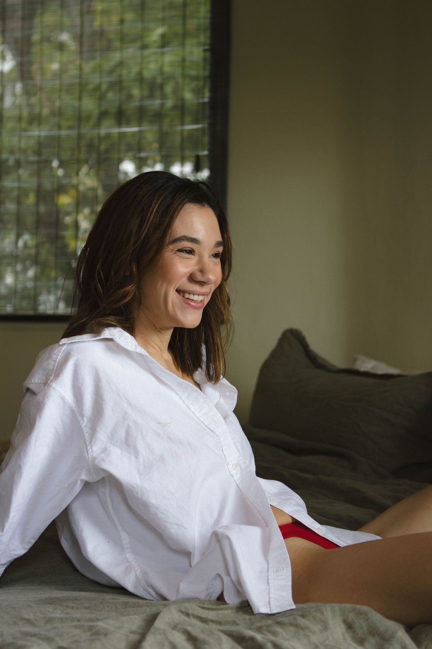A person wearing a white button-down shirt relaxes on a bed with green pillows while smiling.