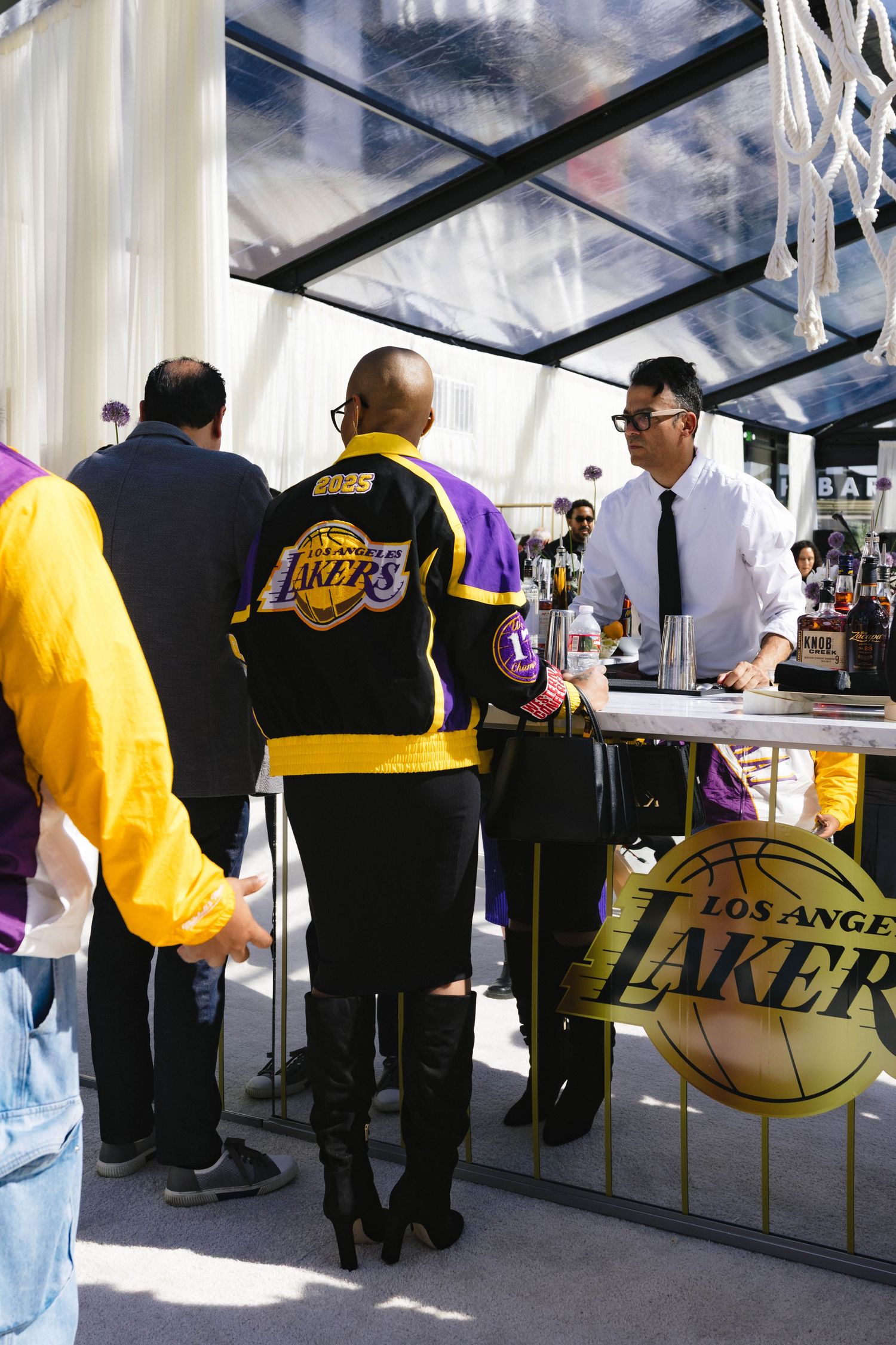 Guests in LA Lakers jerseys at an outdoor Lakers branded bar with glass roof and gold signage.