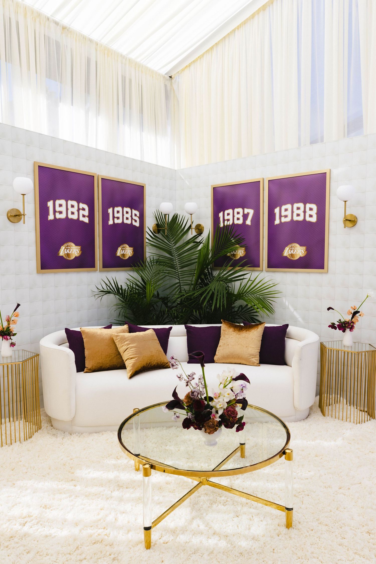 Elegant lounge with white sofa, gold pillows, LA Lakers championship banners from 1982-1988, and a glass coffee table.