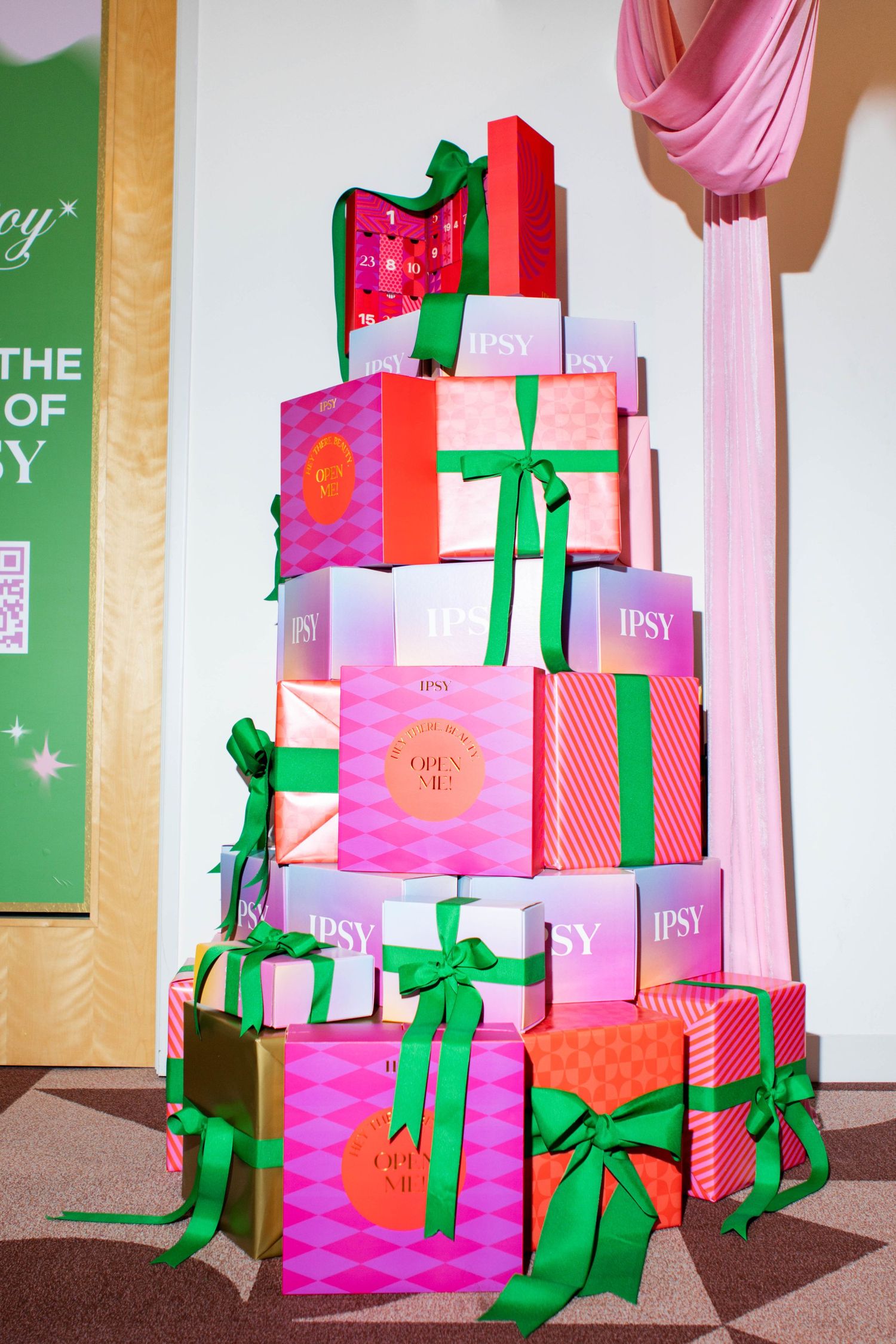 Stack of pink and red Ipsy gift boxes decorated with green ribbons arranged in festive tower display.