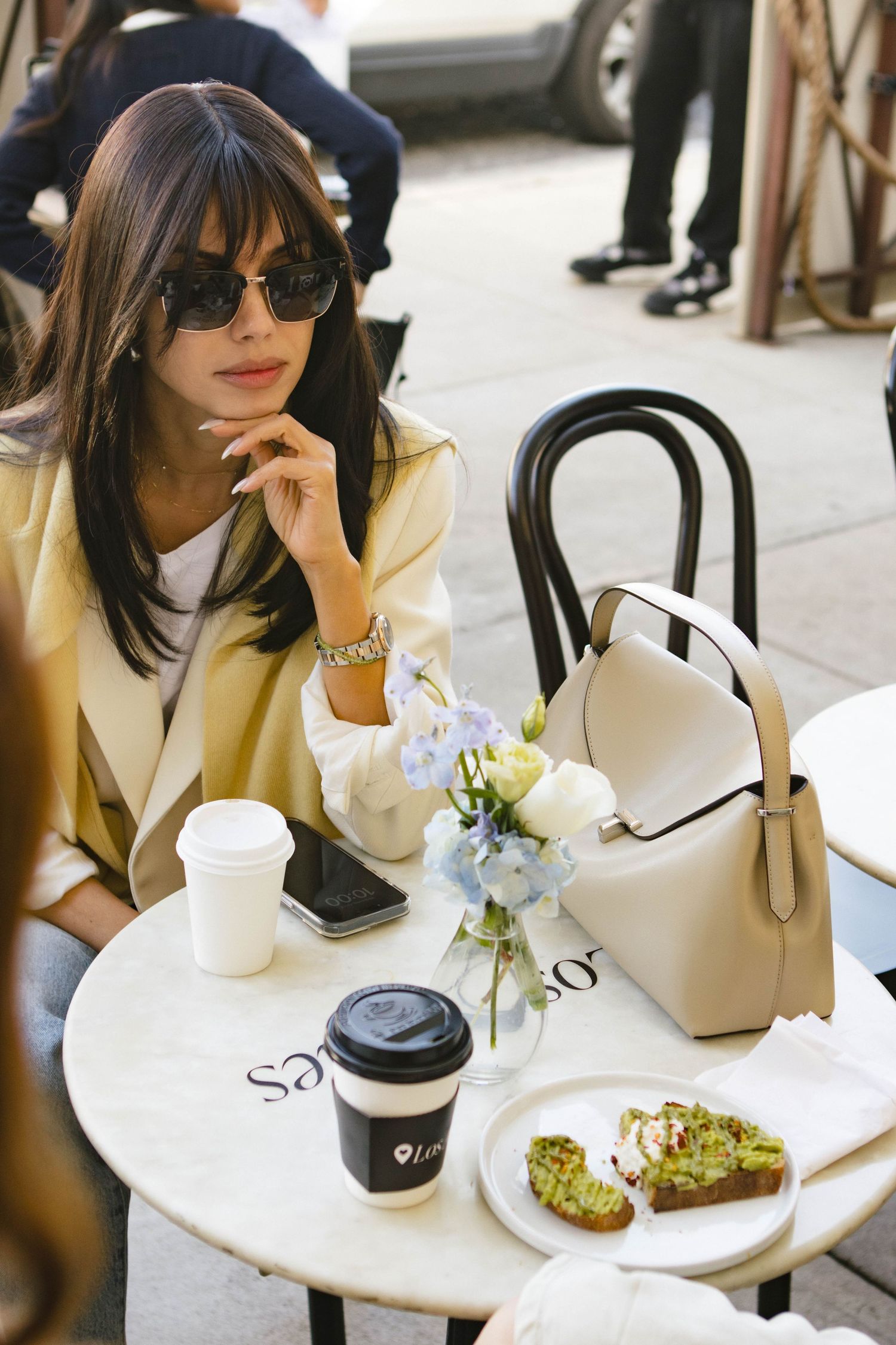 Casual outdoor cafe scene with avocado toast, coffee, and handbag on white table.