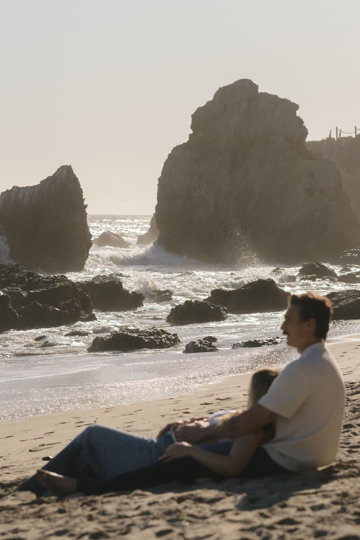 Couple relaxing on a beach with rocky cliffs and crashing waves in the background.