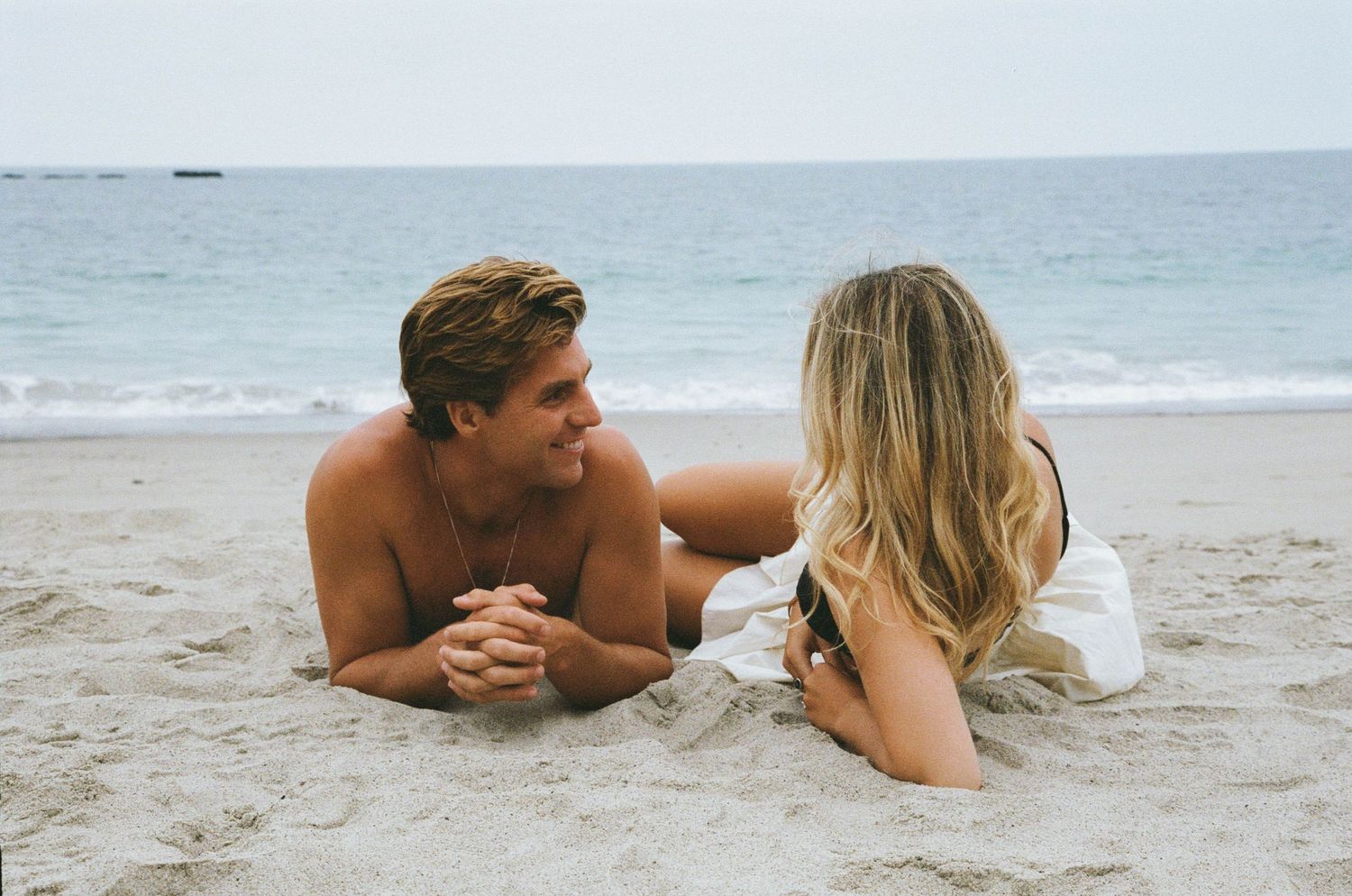 Couple lying on sandy beach with ocean waves in background, captured in romantic seaside photoshoot.