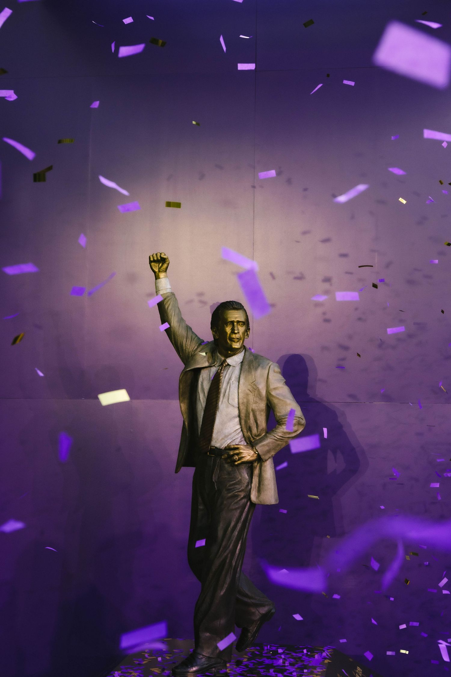 Golden statue of a man with raised fist surrounded by falling purple and gold confetti under dramatic violet lighting.