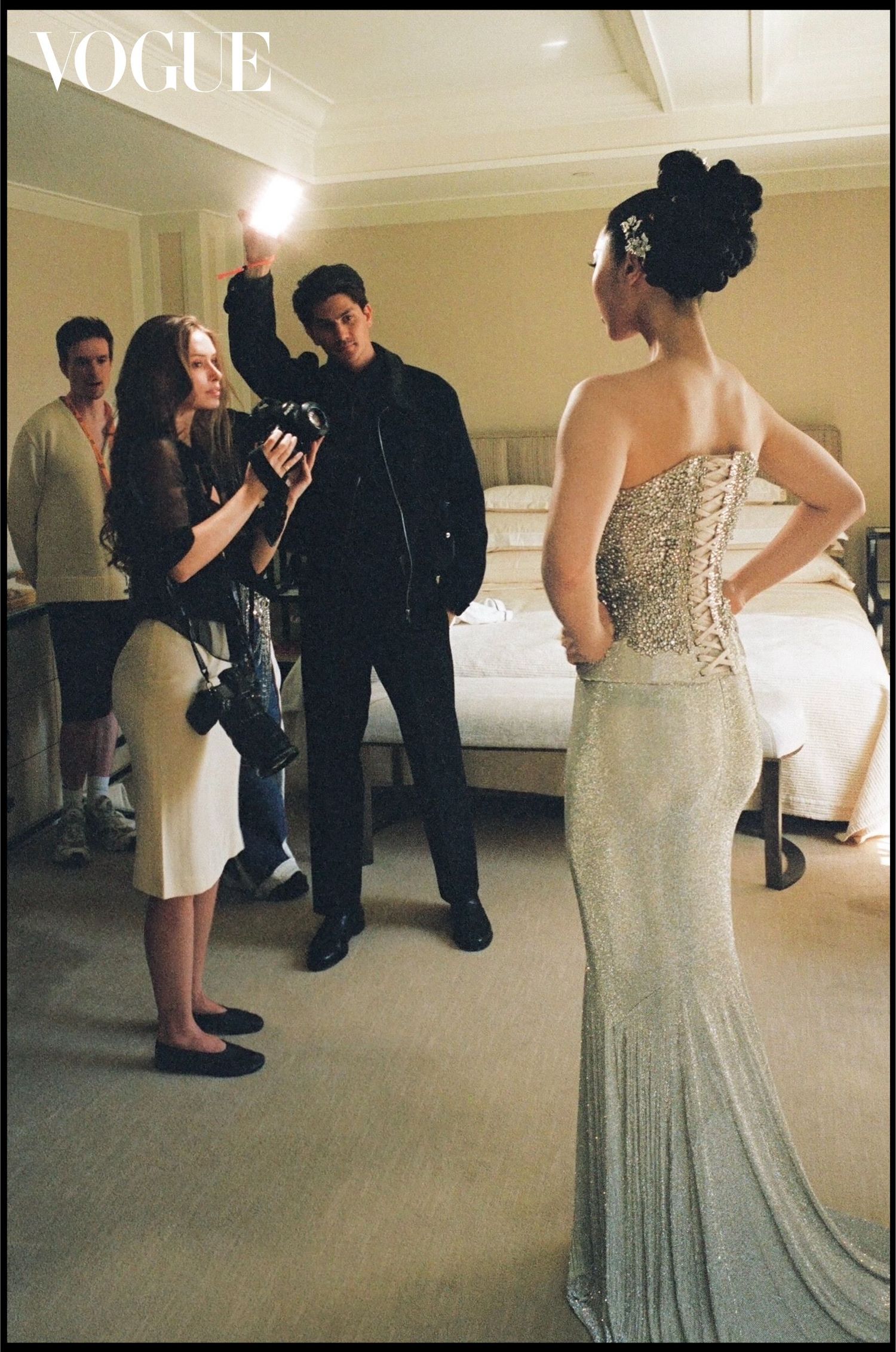 Vogue behind-the-scenes shot of a photographer capturing a woman in a strapless beaded silver gown with an updo as crew members look on.