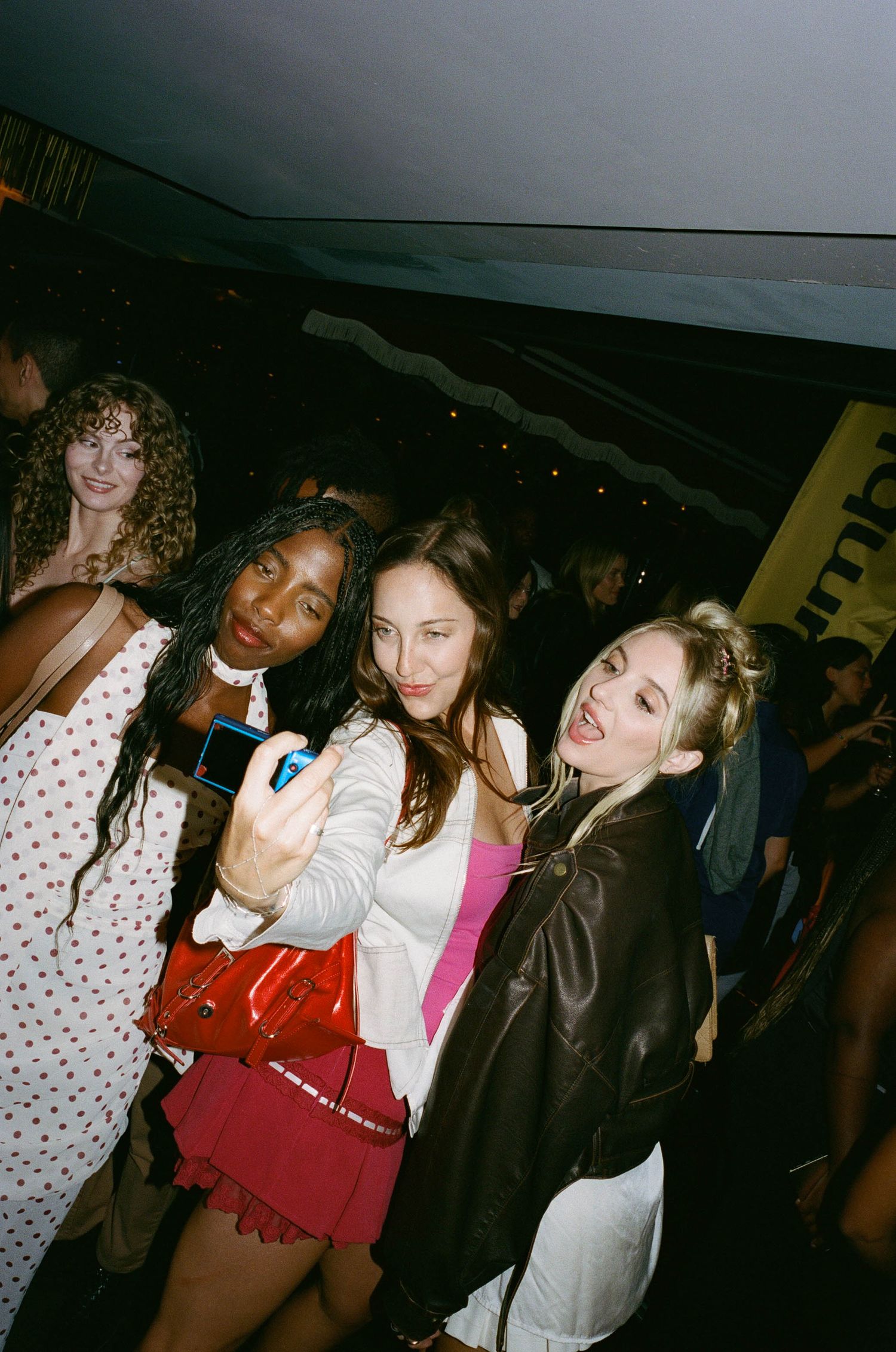 Three young women taking a selfie at a nightclub party, one in a red skirt, one in white, one in a leather jacket.