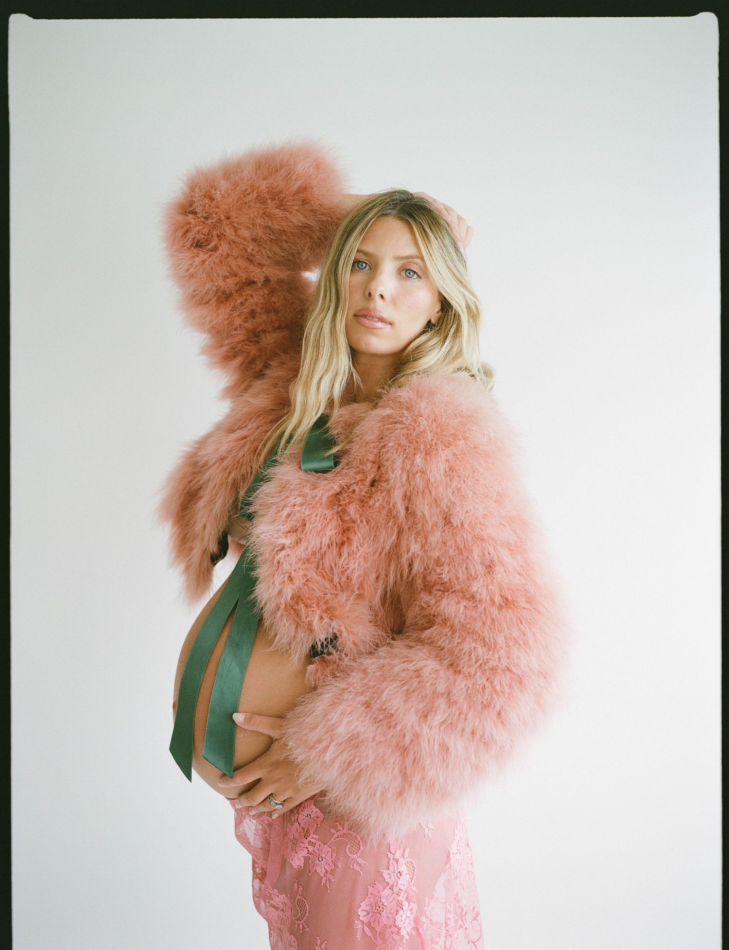 Pregnant woman in pink feather coat and green dress cradling baby bump in a stylish maternity photo shoot.
