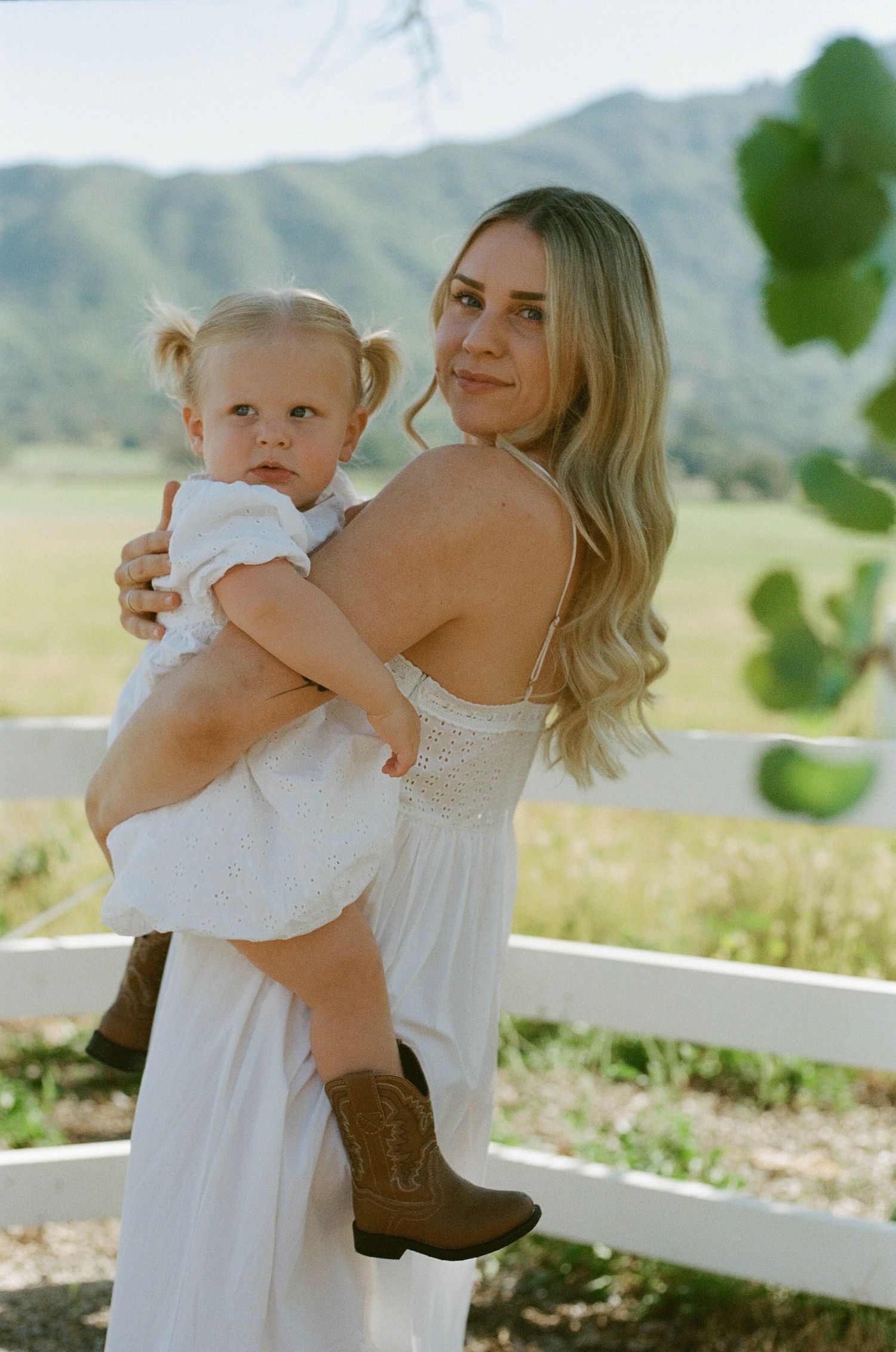 Blonde mother in white sundress holds toddler daughter in cowboy boots with mountain backdrop.