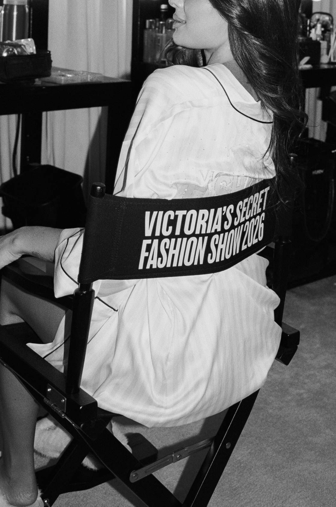 Black and white photo of a woman in a white satin robe seated in a director's chair labeled Victoria's Secret Fashion Show 2026 backstage.