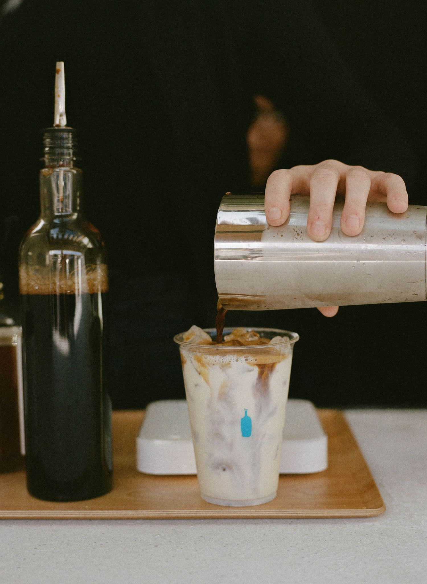 Barista pouring coffee from a shaker into an iced latte cup beside a dark syrup bottle on a wooden tray.