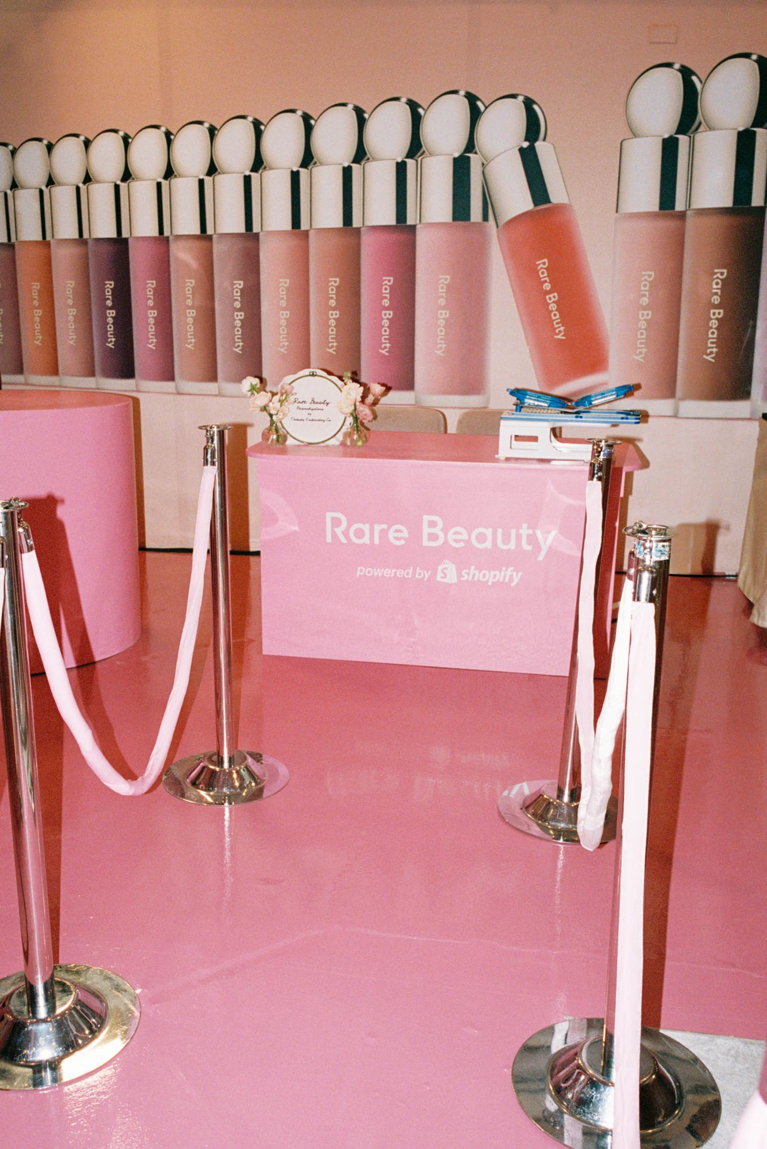 Pink Rare Beauty makeup display with product testers arranged on white shelving against a pink wall.