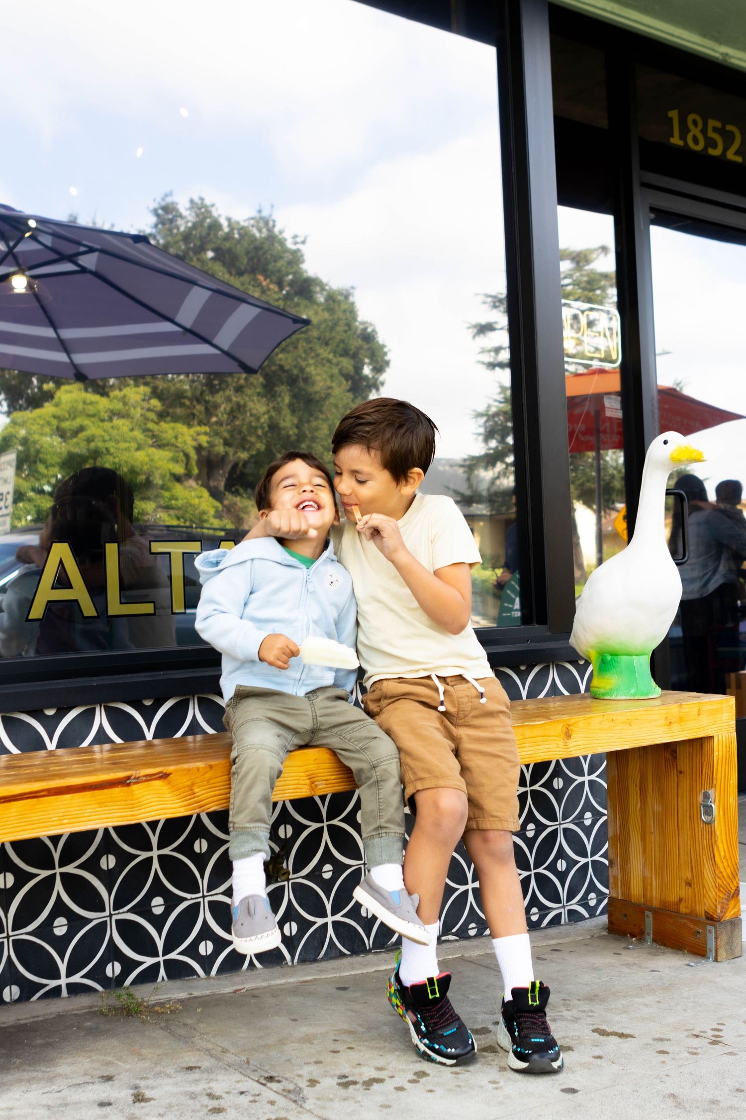 Two children sharing a sweet moment while sitting on a wooden bench outside a storefront.