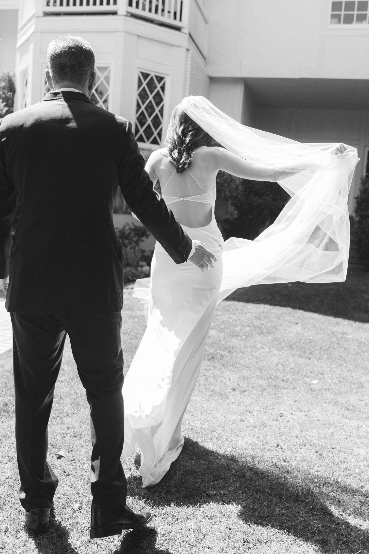 Black and white photo of bride and groom walking hand in hand, her veil flowing in the wind outside elegant venue.