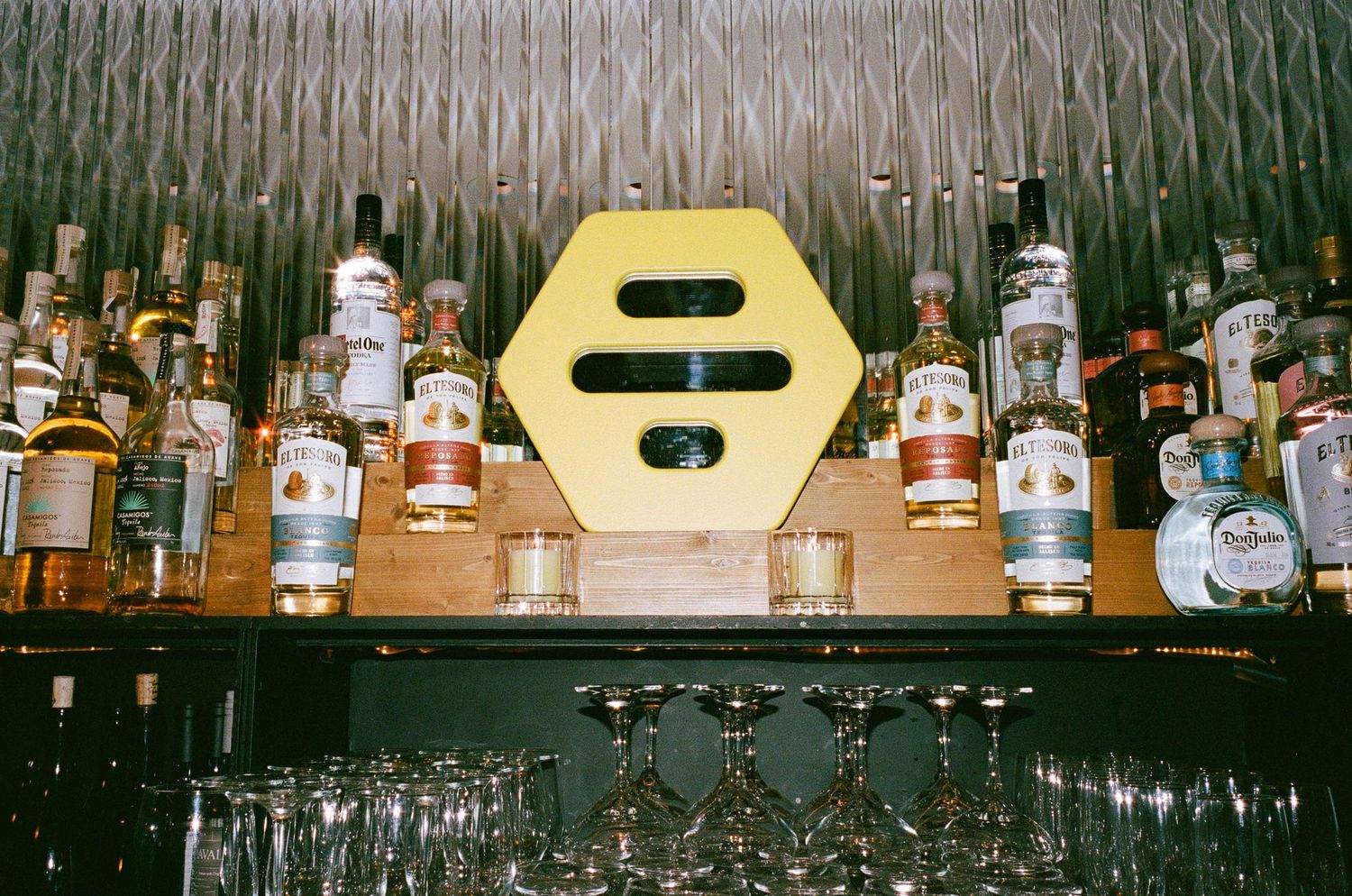 A well-stocked bar shelf displaying various liquor bottles behind a yellow hexagon sign with wine glasses below.
