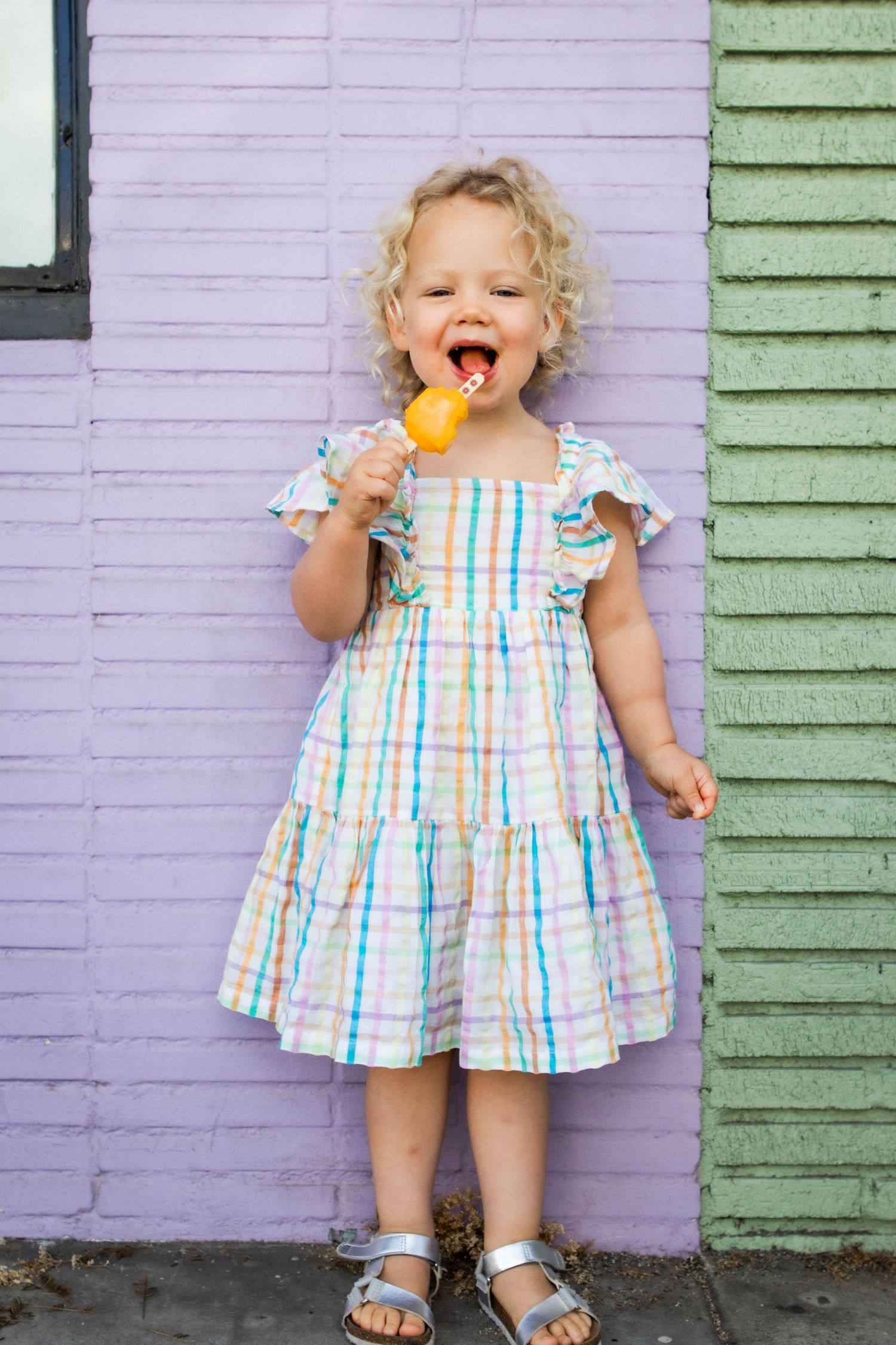 A toddler in a striped summer dress enjoys a yellow popsicle against a purple and green wall.