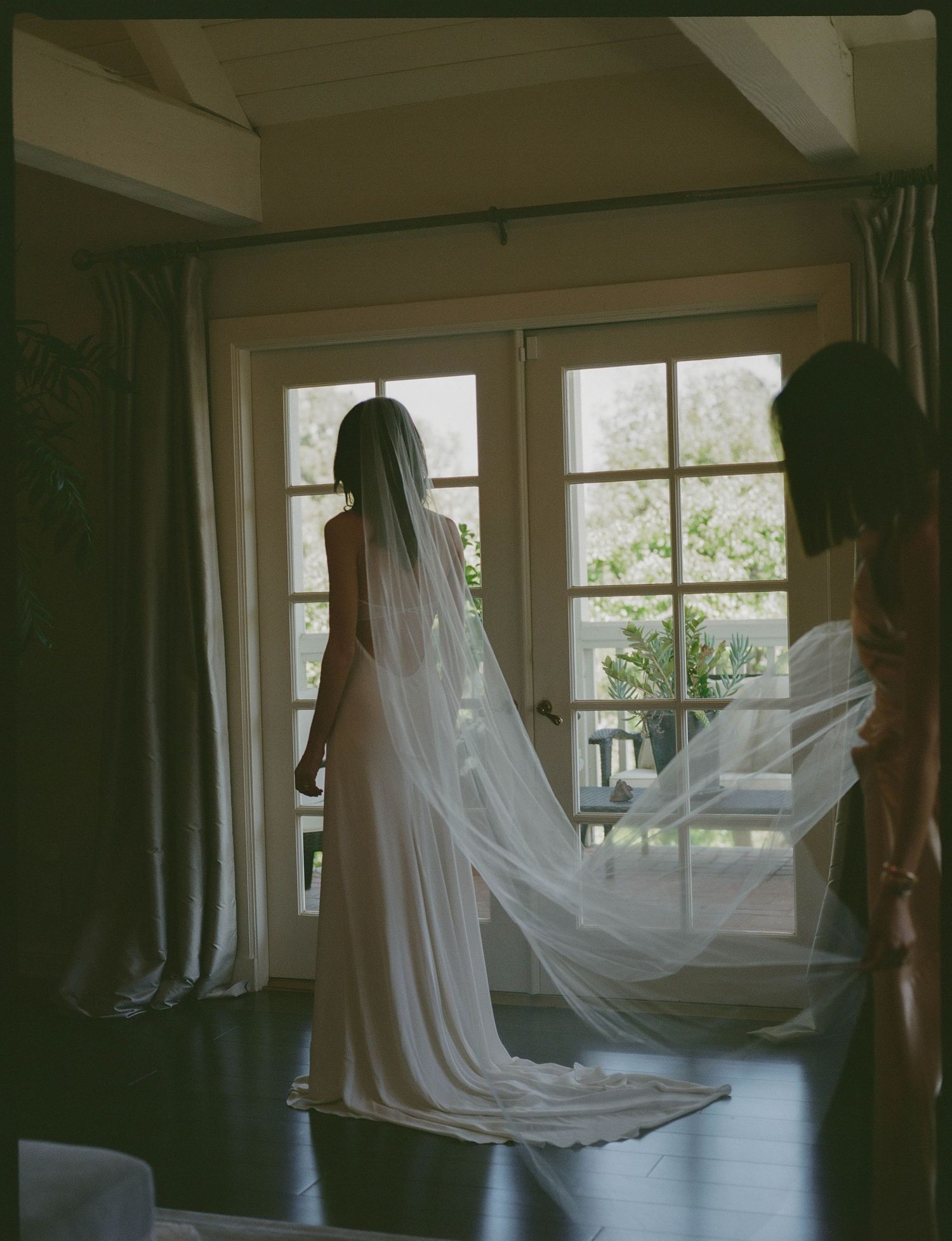 Bride in elegant white gown and long cathedral veil stands by French doors while bridesmaid holds veil train.