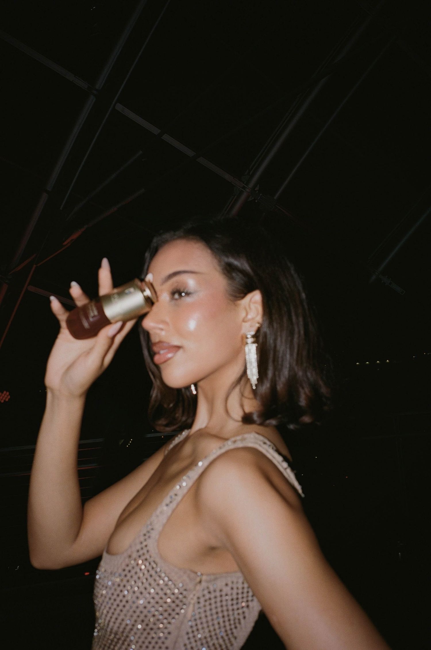 Woman holding a Clarins beauty product near her face, wearing a sparkly dress and chandelier earrings.