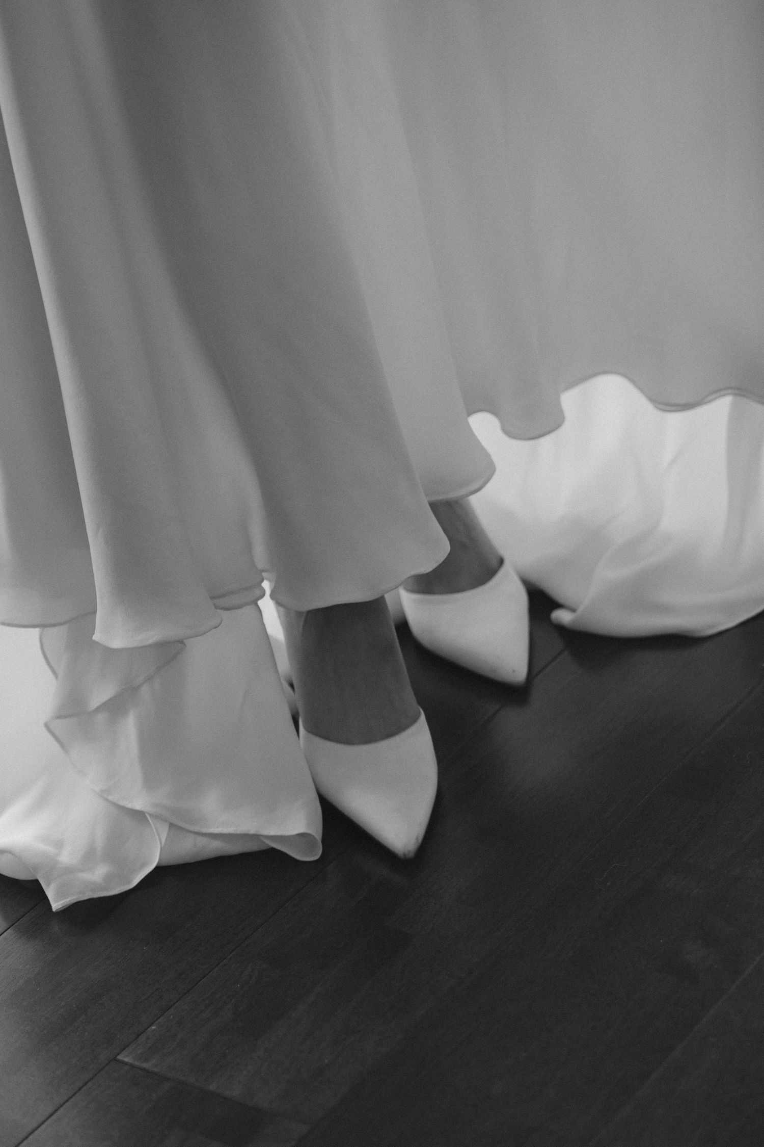 Black and white photo of a bride wearing white pointed heels and a flowing layered wedding gown on dark hardwood floors.